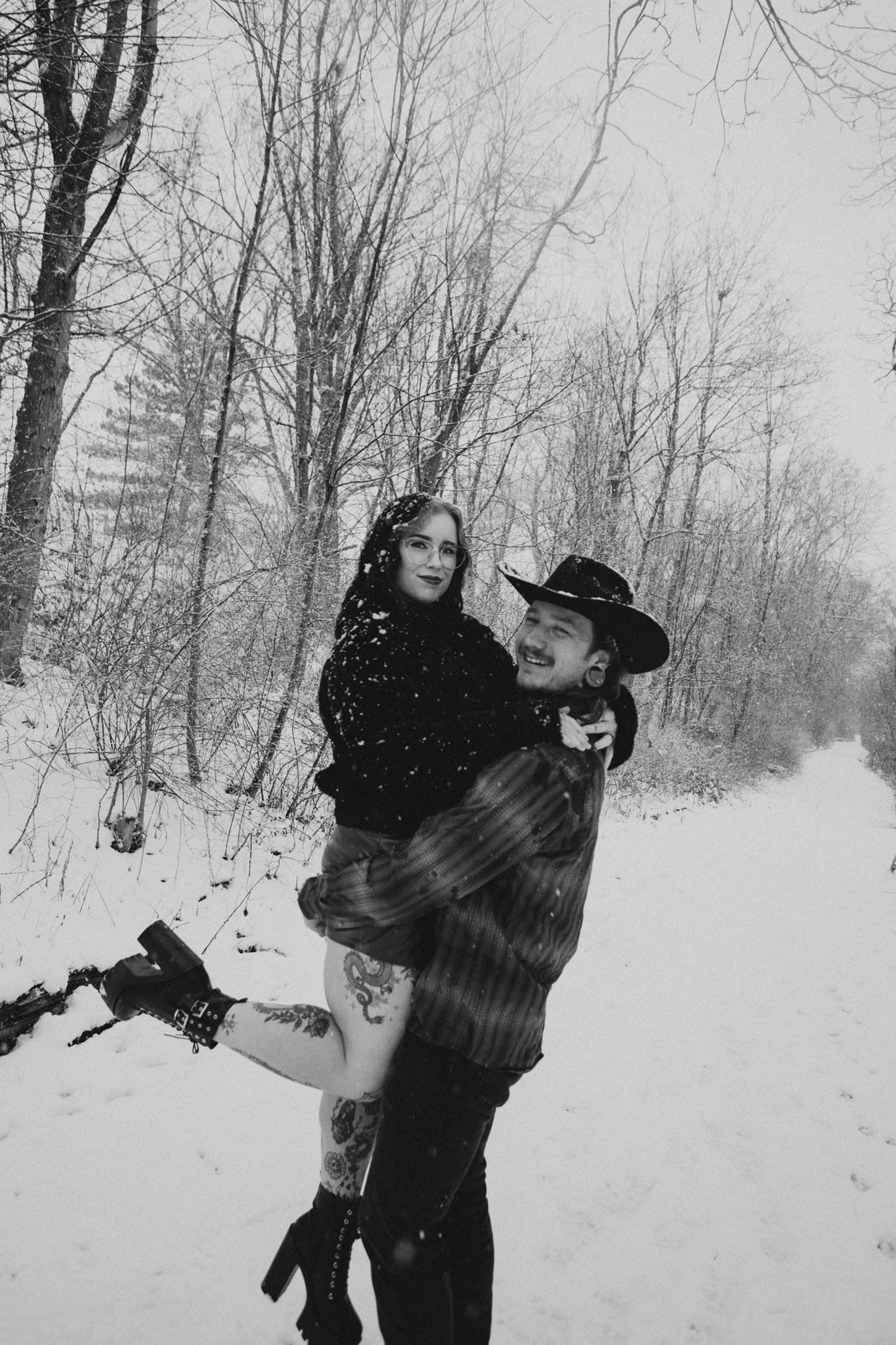 A man holding a woman in a snowy forest, both smiling, with the woman wearing glasses and boots and the man wearing a hat.