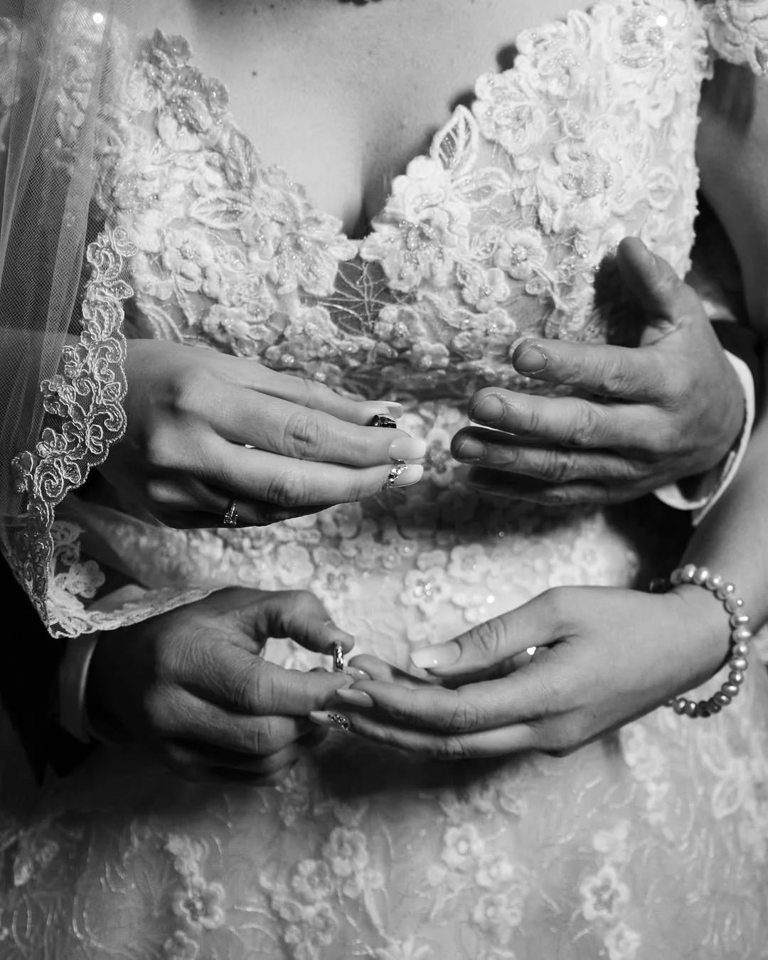 Close-up of a bride and groom holding hands, with the bride wearing an embroidered wedding dress and the groom in a suit. Their wedding rings are visible.