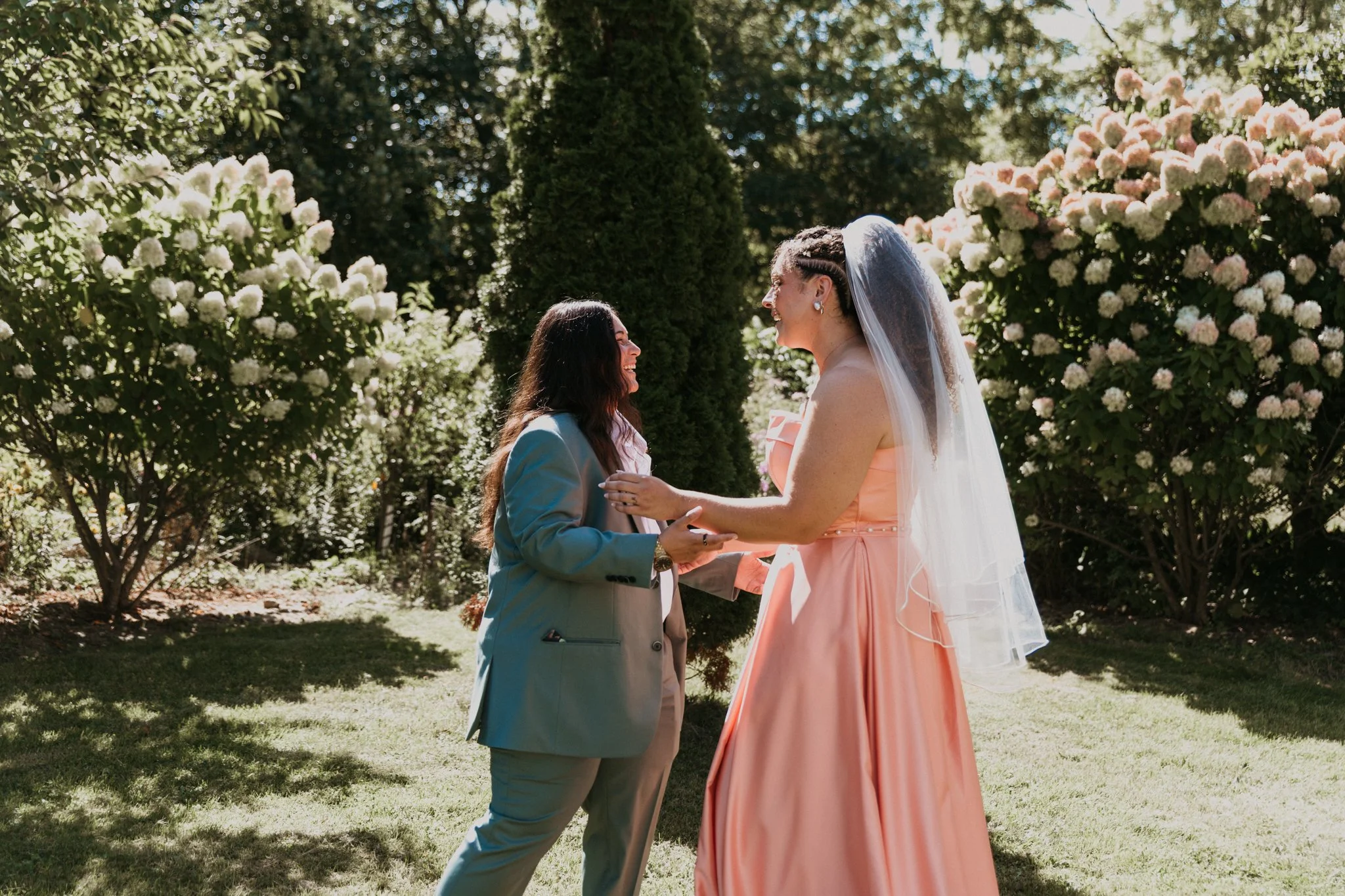 Two women hold hands and smile at each other during a wedding ceremony outdoors, surrounded by lush greenery and blooming bushes.