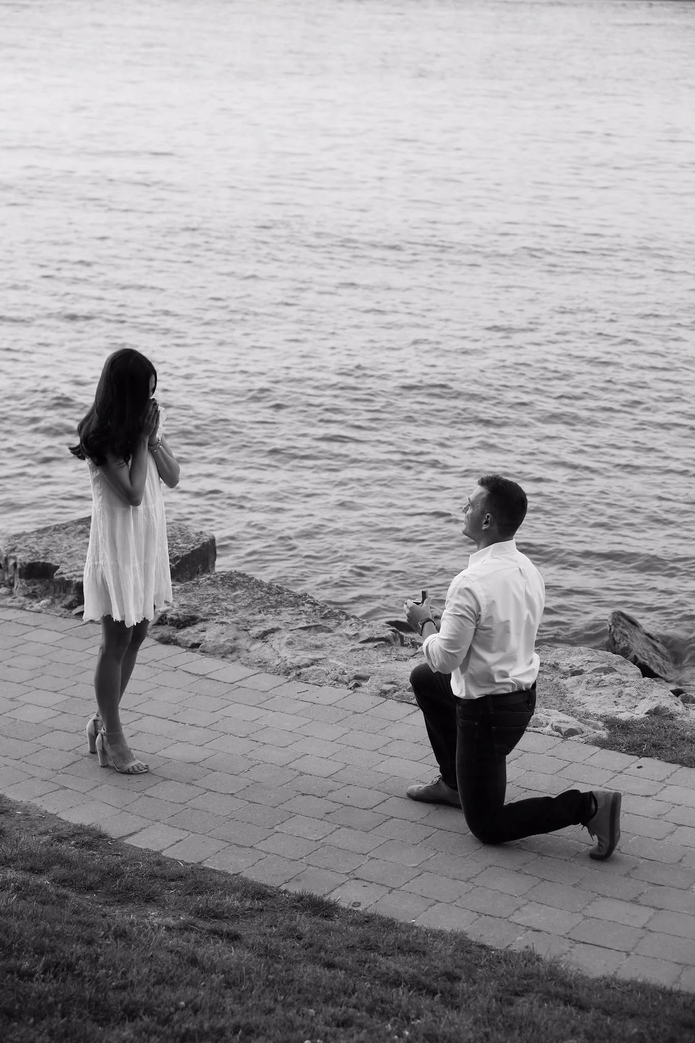 A black and white photo of a man proposing marriage to a woman by the water, with the man kneeling on one knee and the woman covering her mouth in surprise.