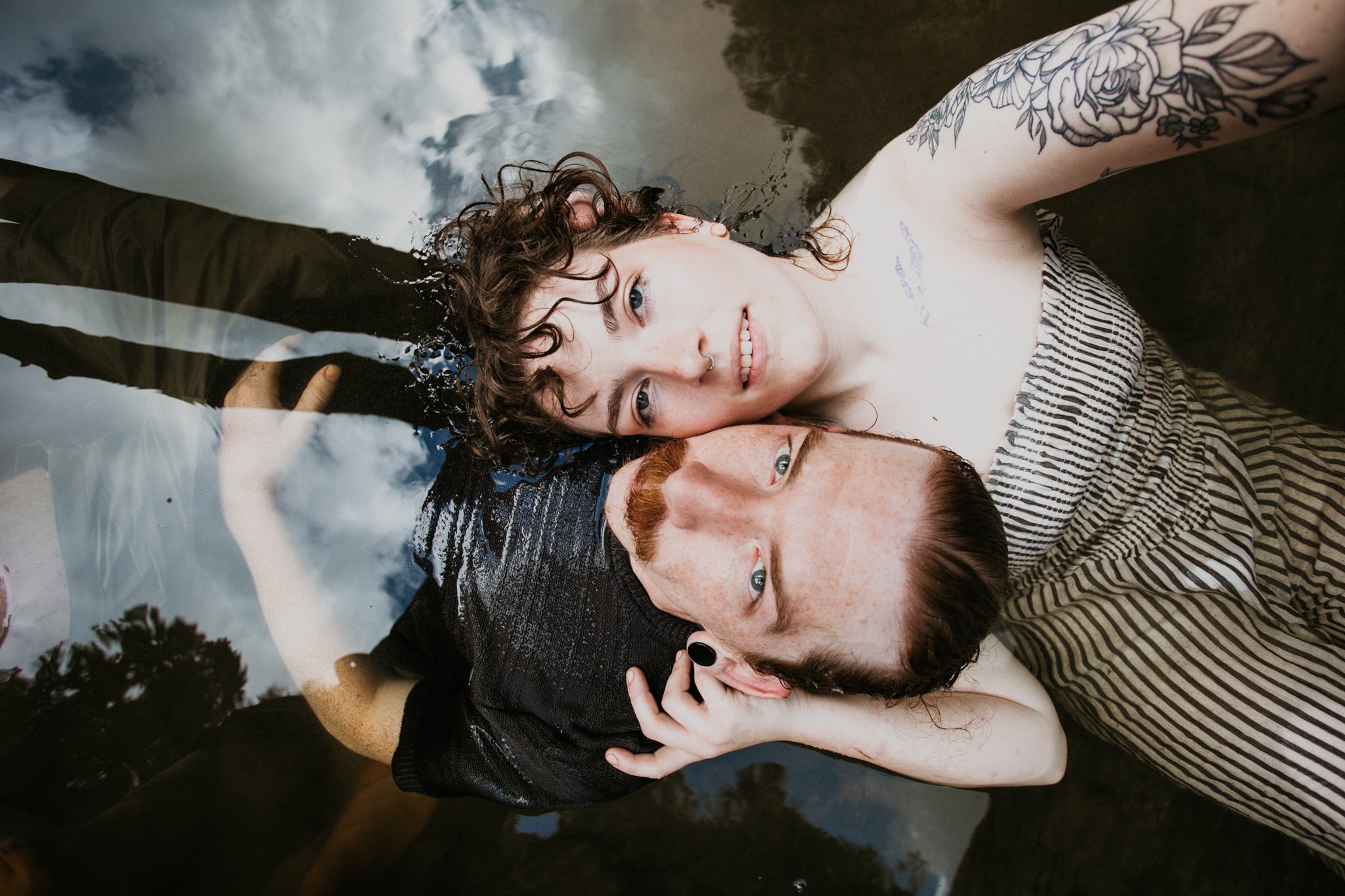 Two young adults, a woman with curly brown hair and a man with red hair and a mustache, are lying in shallow water outdoors, looking upward and smiling. The reflection of the sky and trees is visible in the water around them.