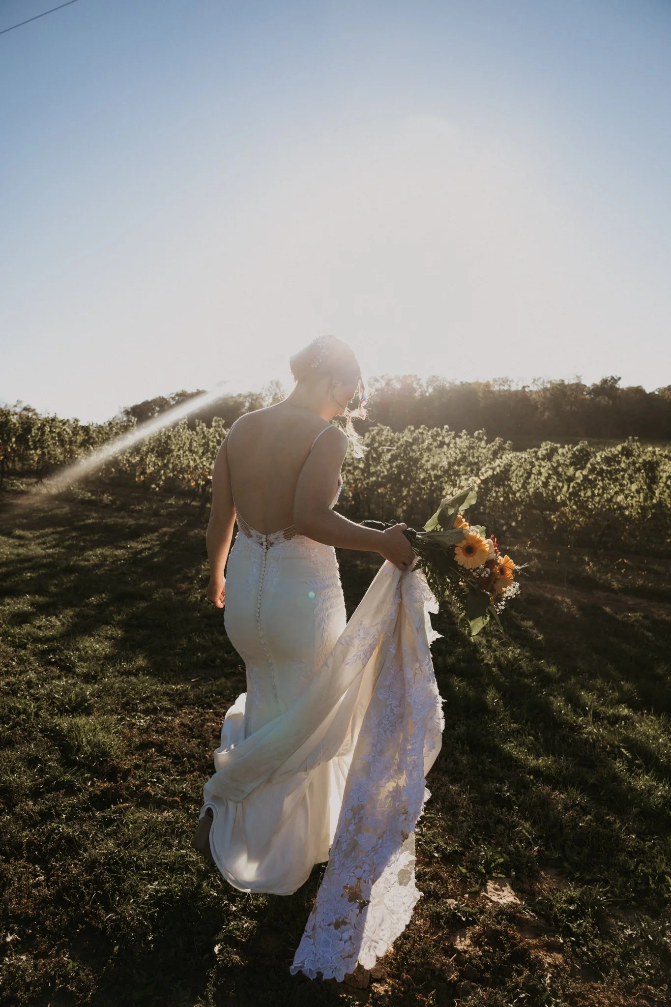 A bride in a white wedding dress holding a bouquet of flowers, walking in a sunny field with the sun shining brightly behind her.