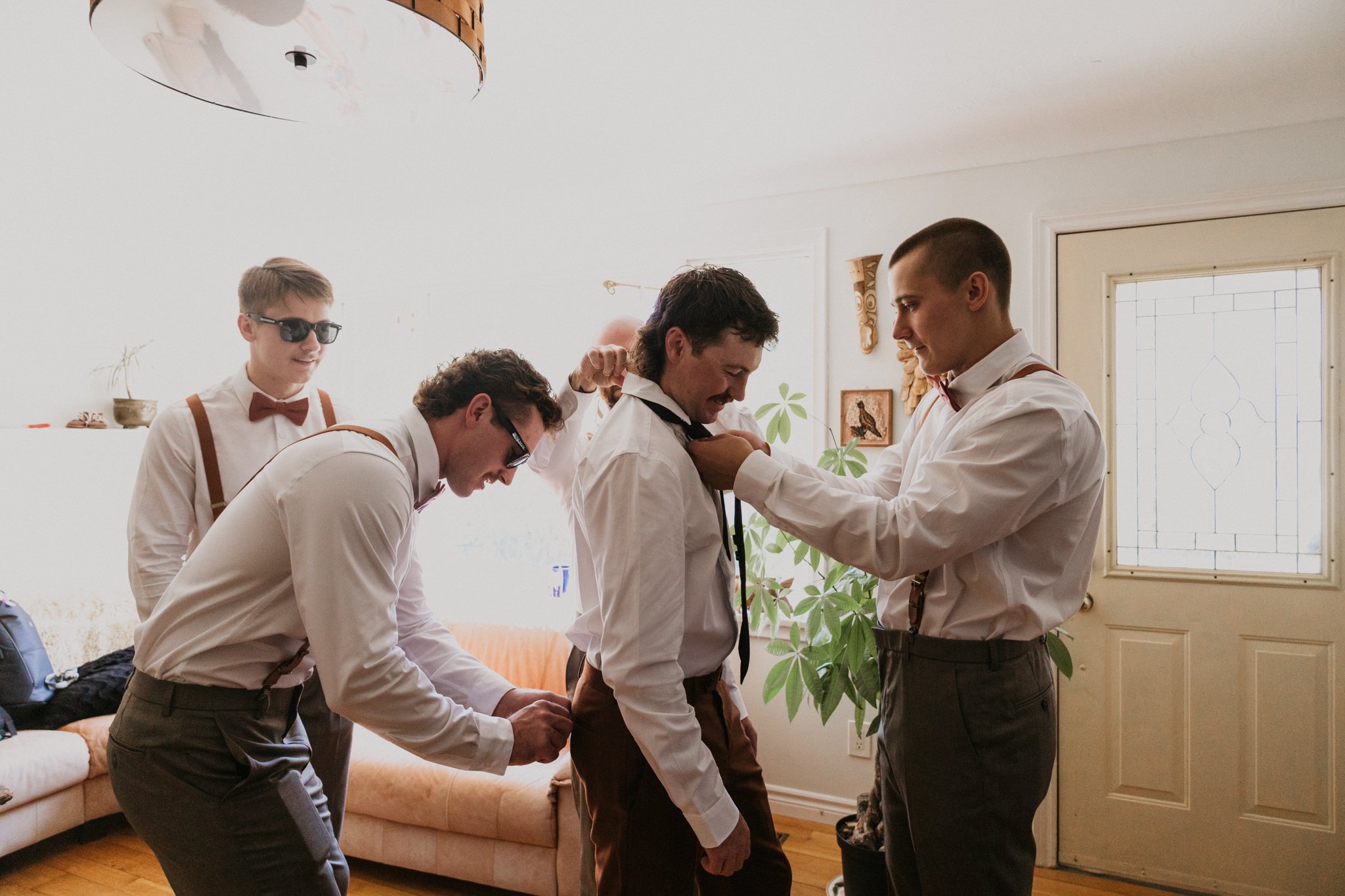 Group of men getting dressed up, with one man adjusting his tie while others assist, in a well-lit living room with a couch, plants, and wall decorations.