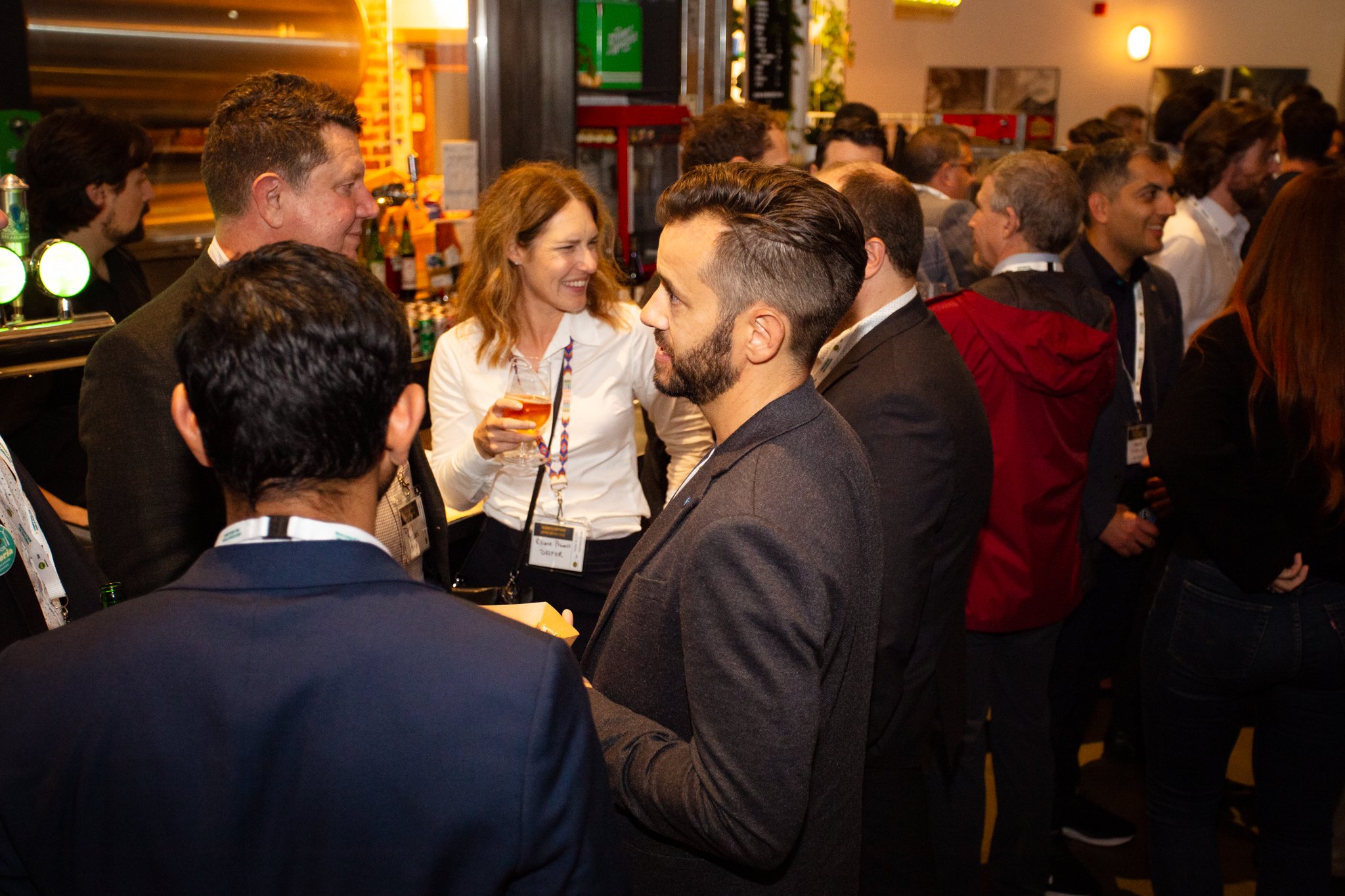 Group of people at a networking event, talking and holding drinks, in a lively indoor venue with bar and industrial design elements.