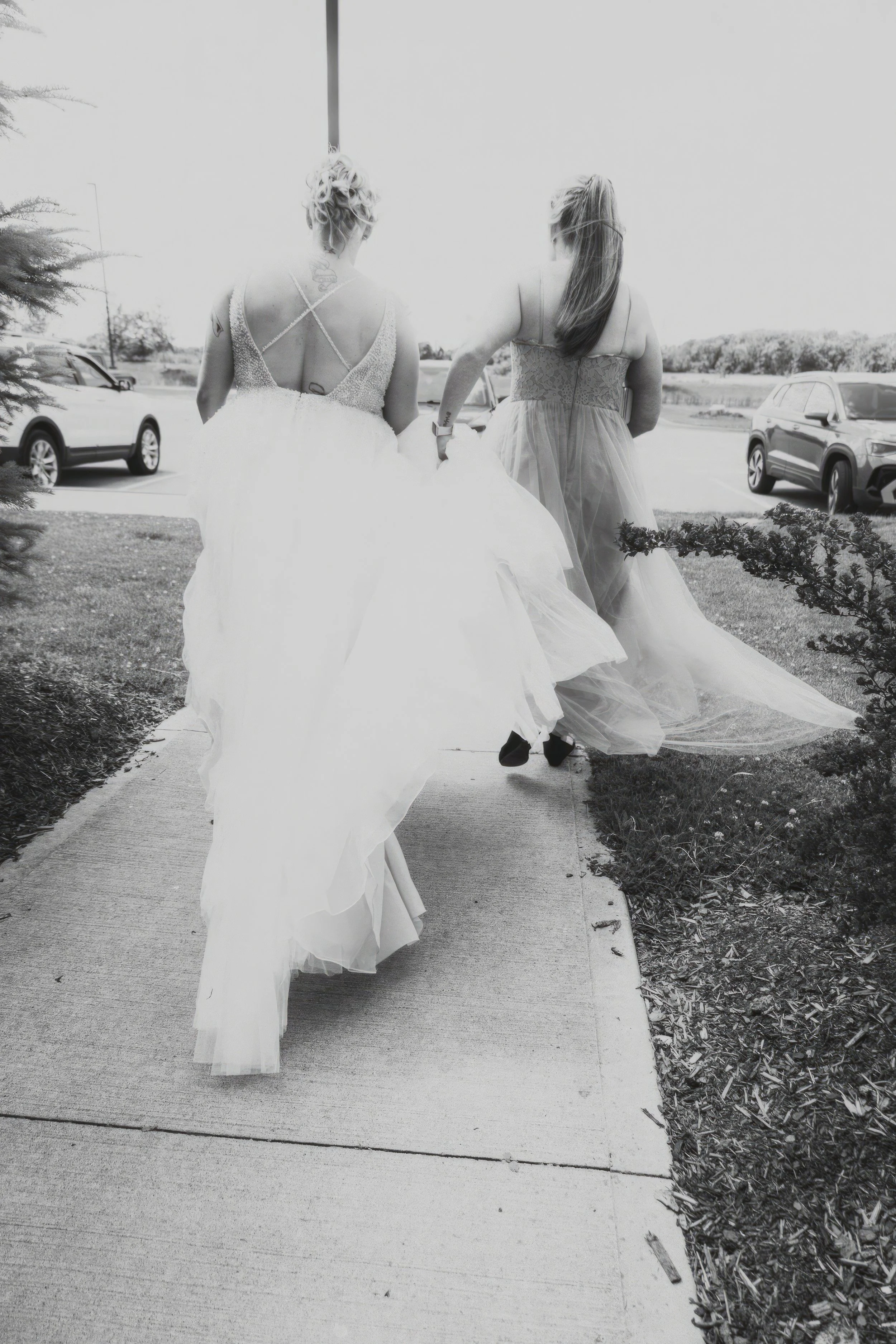 Two women in wedding dresses walking on a sidewalk outside.