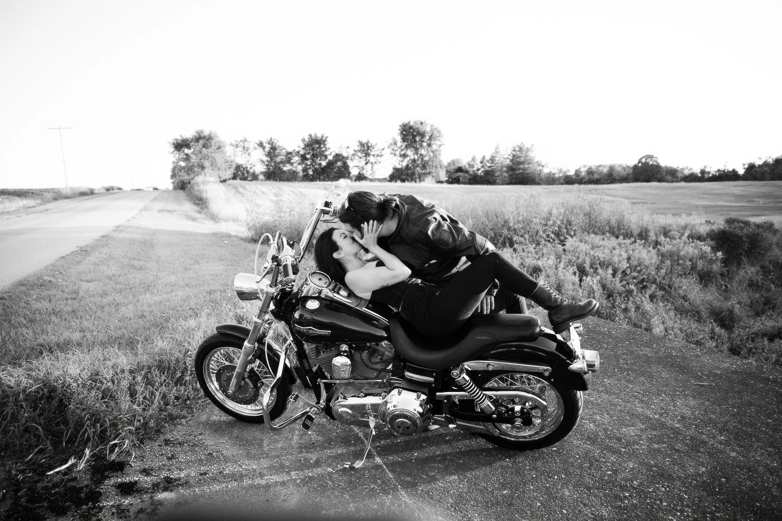 Couple sharing a kiss on a motorcycle by a rural road in black-and-white.