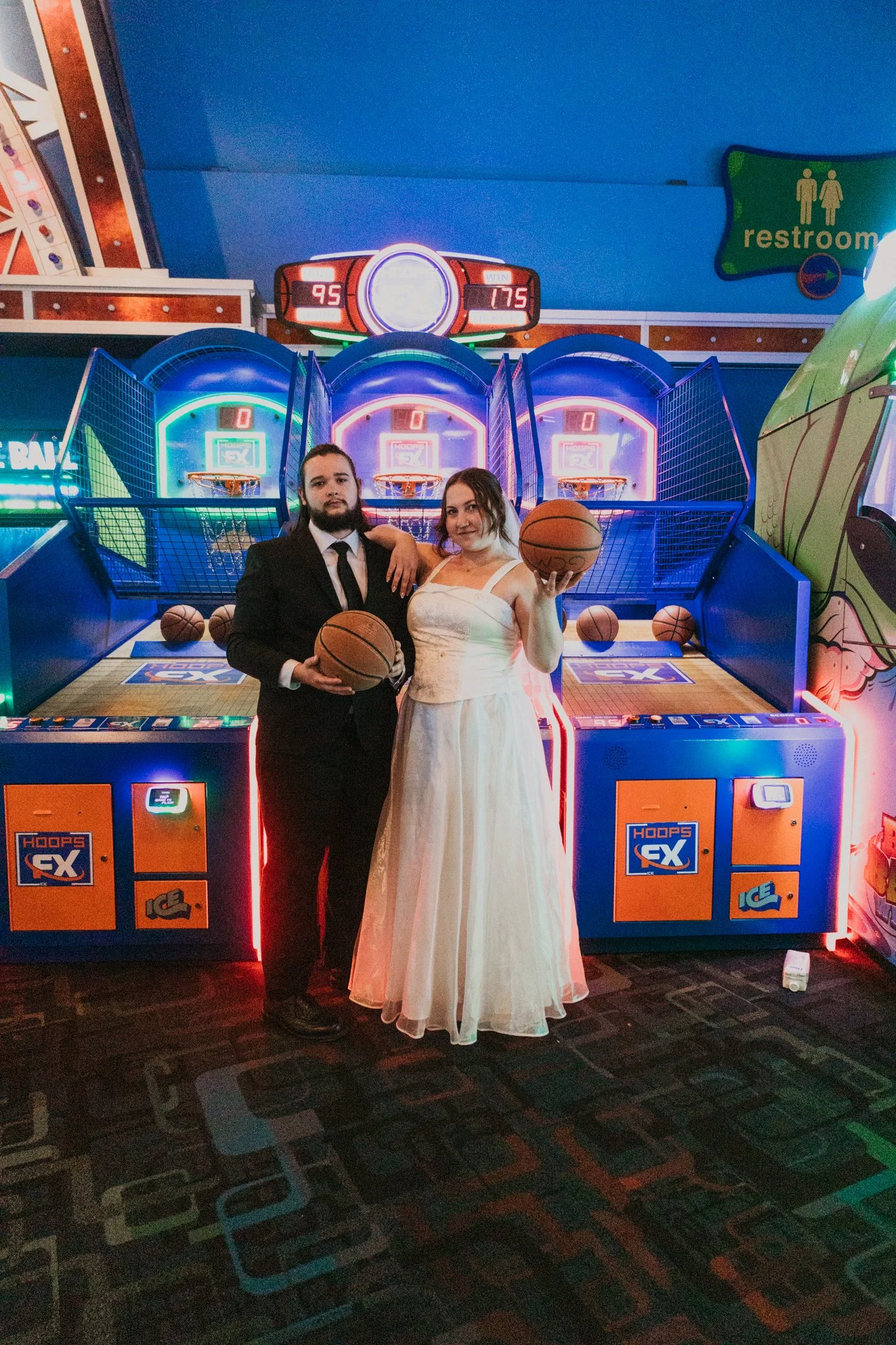 A man in a suit and a woman in a white dress holding basketballs at an arcade basketball game.