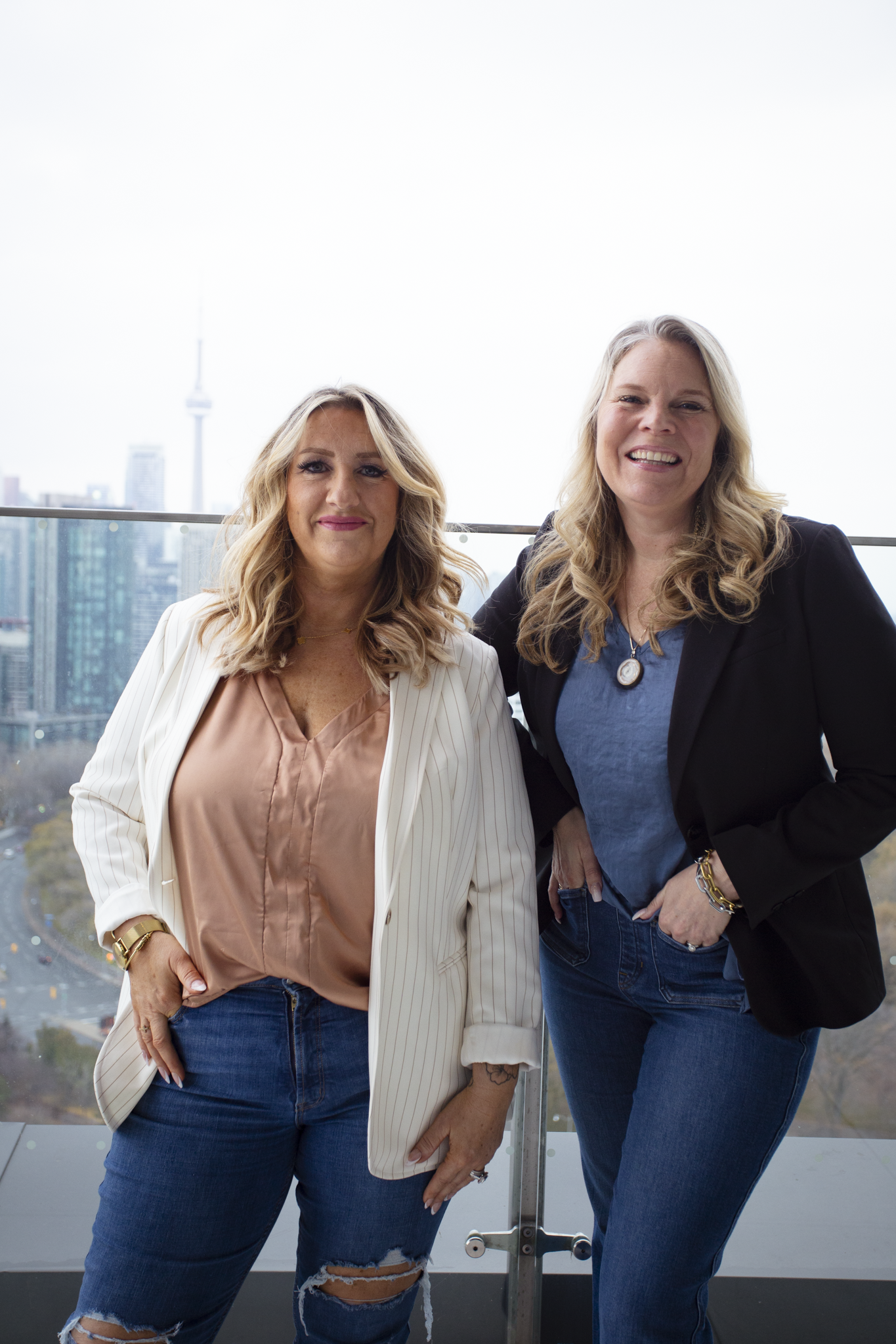 Two women smiling and standing on a balcony with city buildings and the CN Tower in the background.