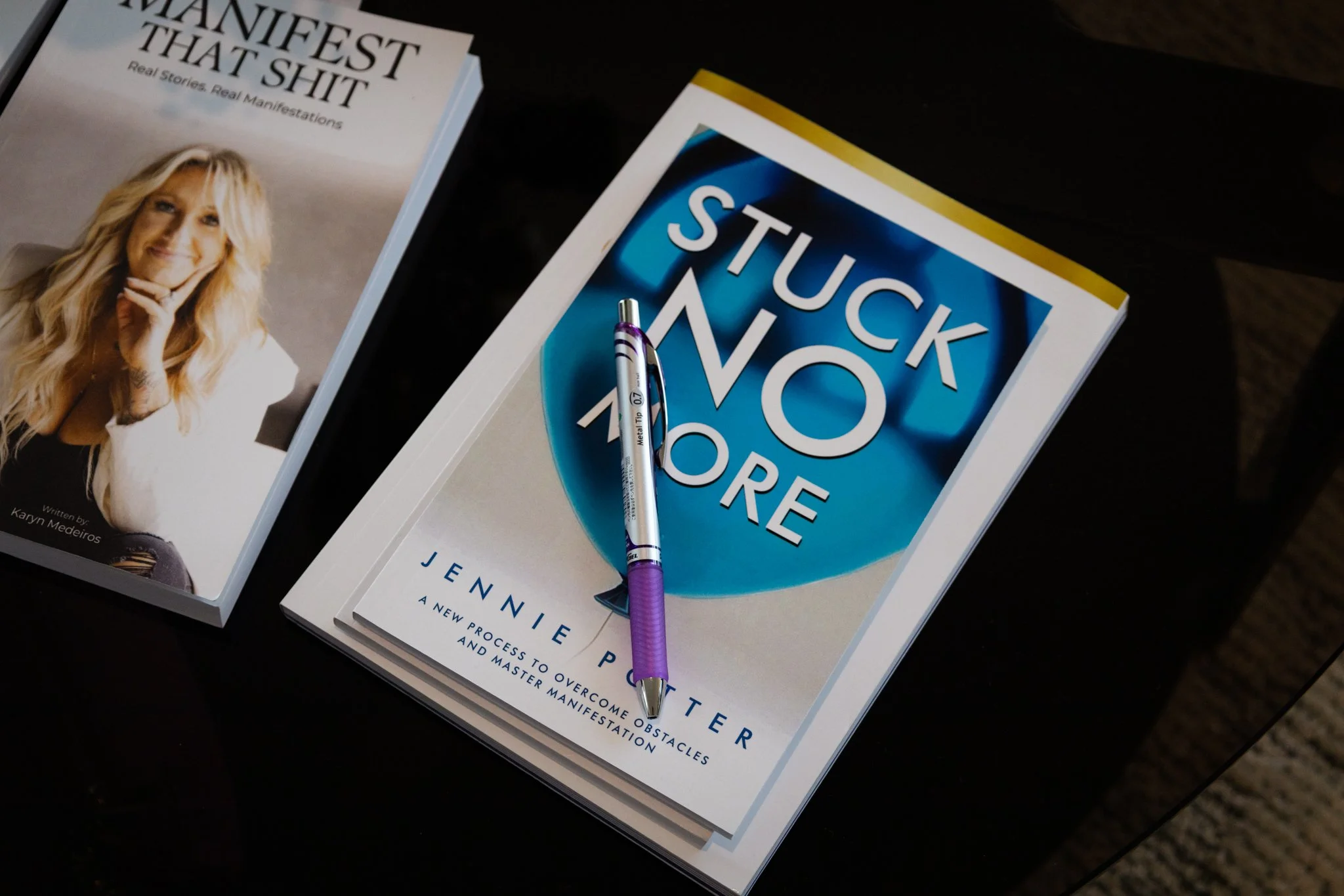A book titled 'Stuck No More' by Jennie Potte, with a purple and white pen resting on it, and a second book titled 'Manifest That Shit' with a picture of the author on the cover.