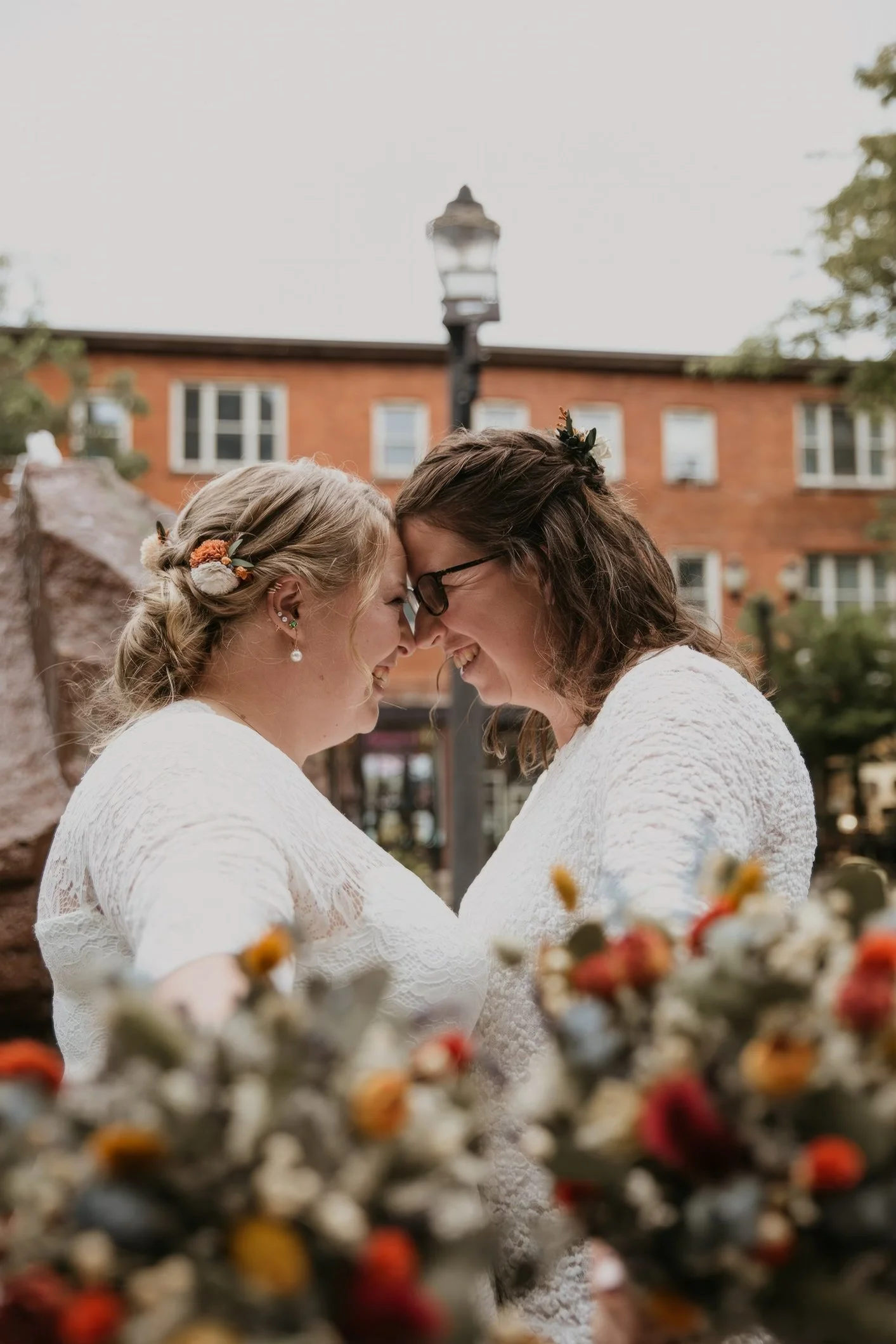 Two women in white dresses about to kiss, smiling, outdoors with a blurred brick building and flowers in the foreground.