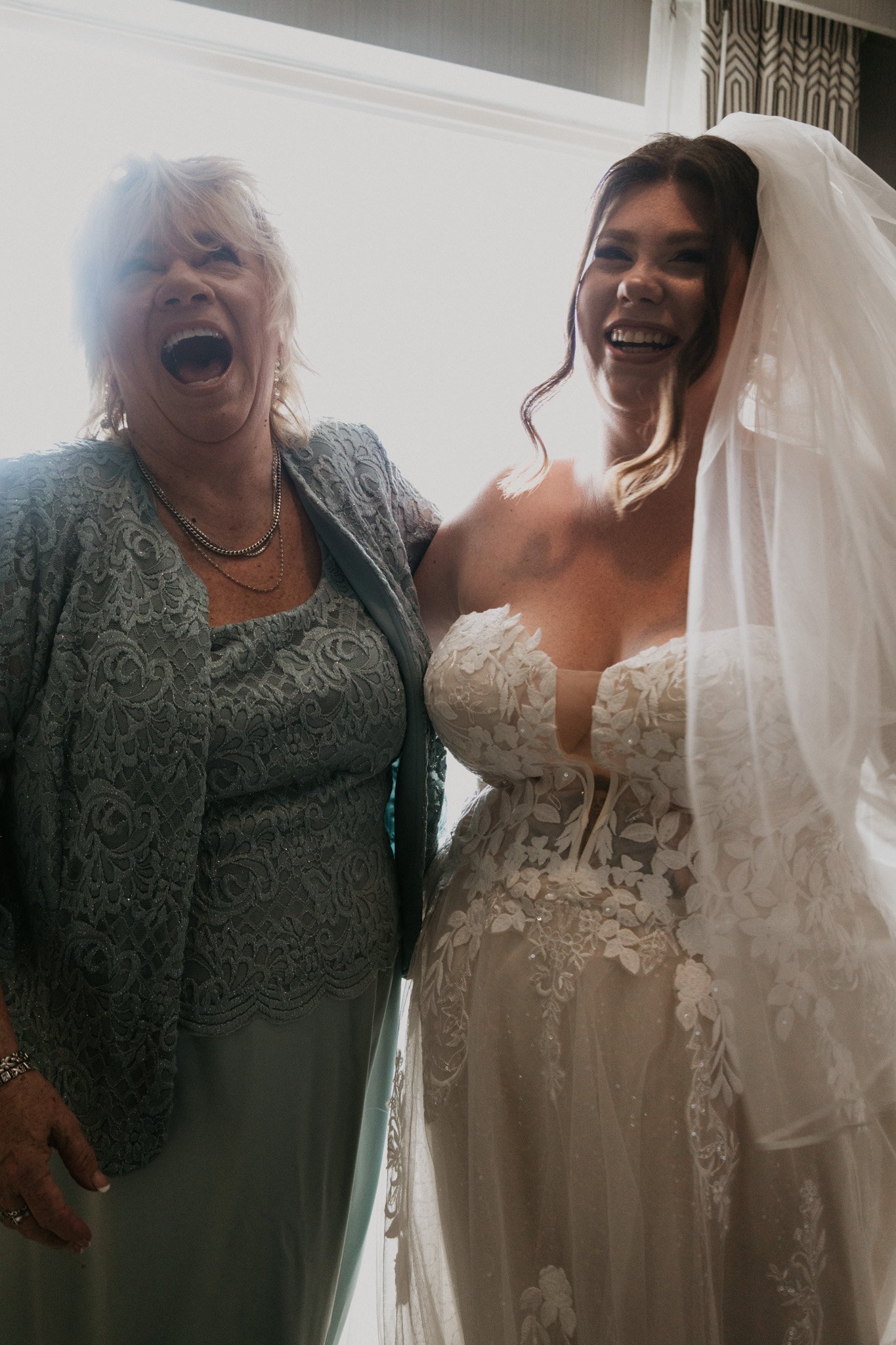 Joyful moment of a bride in a lace wedding dress and veil, and an older woman in a lace dress, sharing a happy laugh indoors with bright natural light behind them.