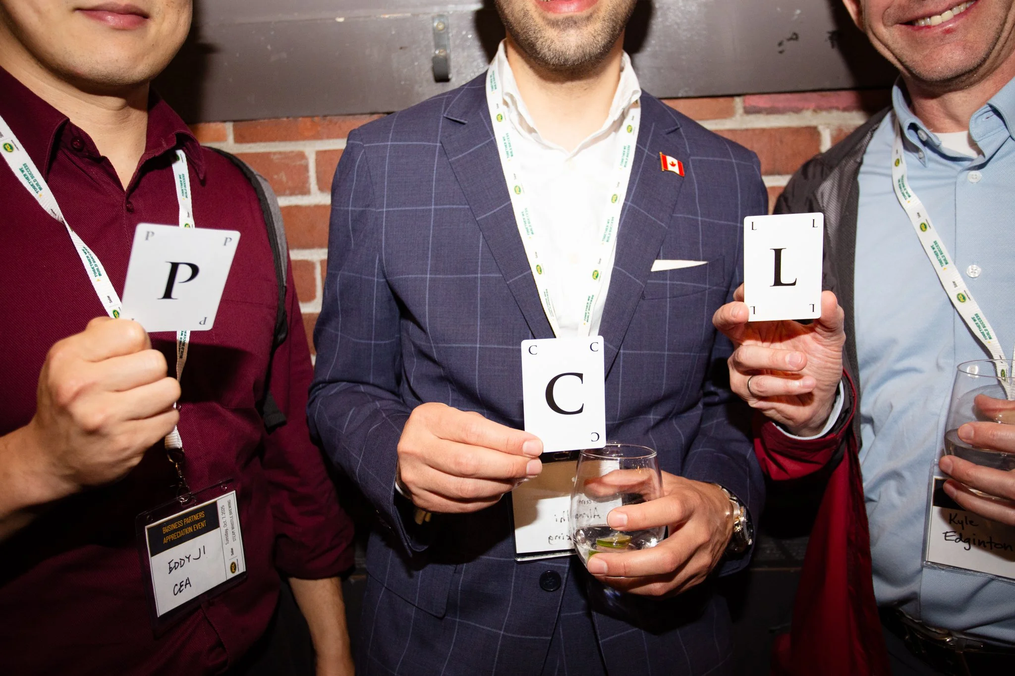 Three men at a networking event holding large alphabet cards with the letters 'P', 'C', and 'L', standing in front of a brick wall.