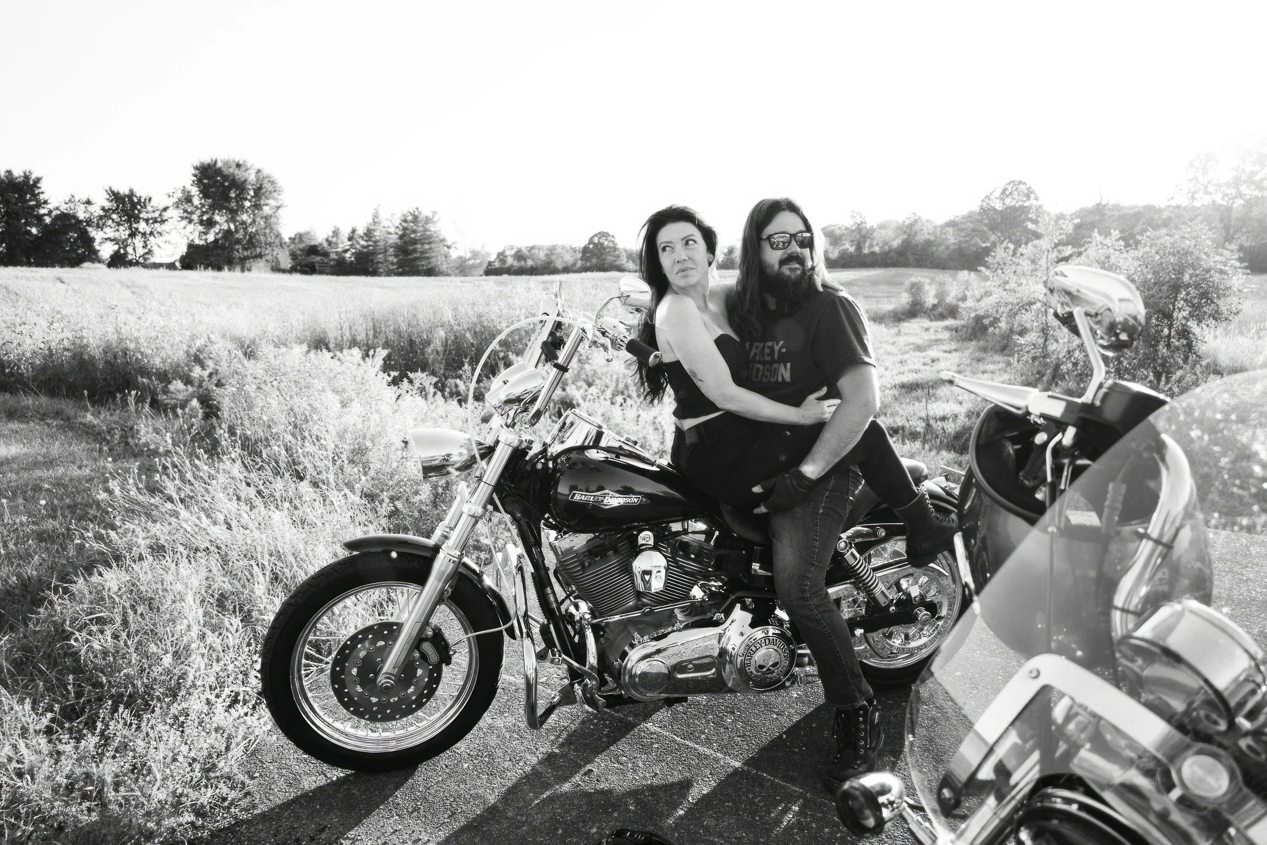 A man and woman sitting on a Harley-Davidson motorcycle in a rural field with trees in the background, both looking off into the distance, black-and-white image.