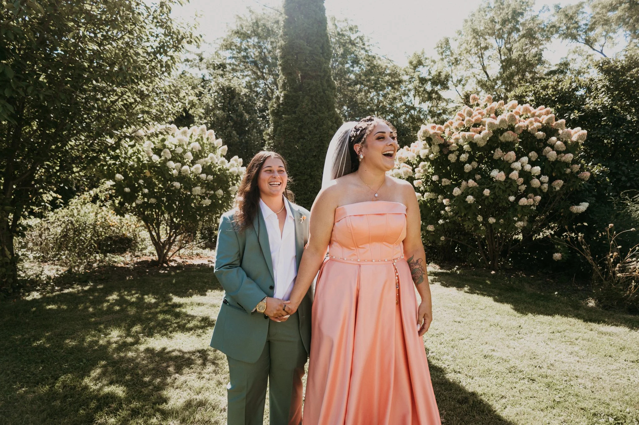 Two women holding hands and smiling outdoors, surrounded by lush green trees and blooming white flowers, during a sunny day.
