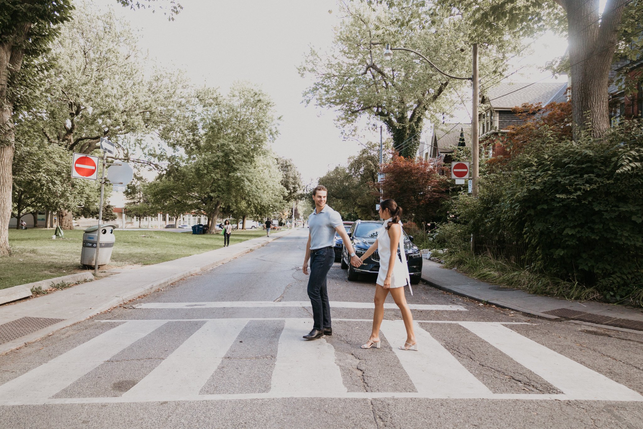 A couple holding hands and crossing a street on a crosswalk, with cars parked along the curb, large trees, and residential houses in the background.