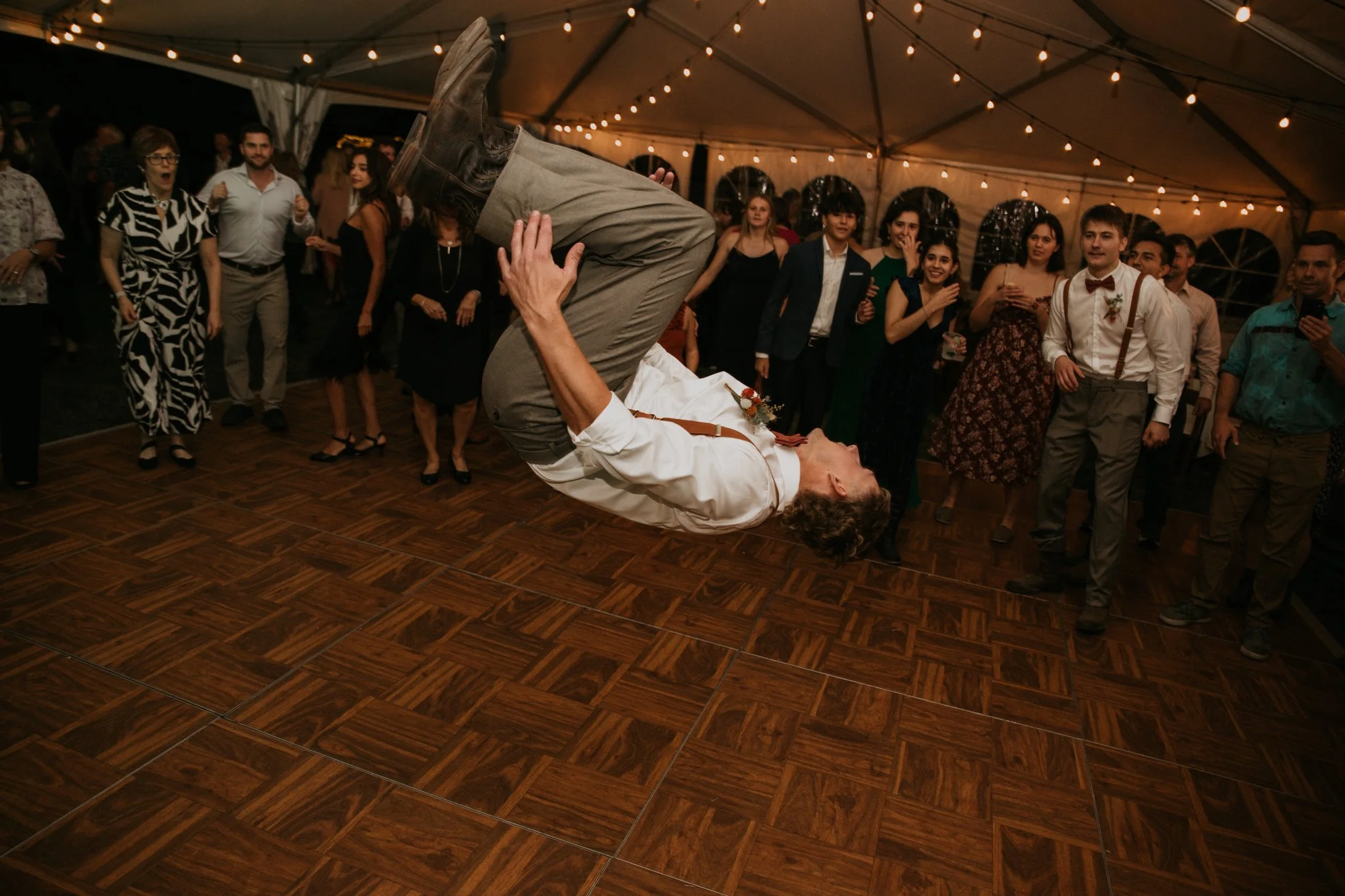 A man is mid-air flipping on a dance floor at a wedding reception, surrounded by guests under a tent with string lights.