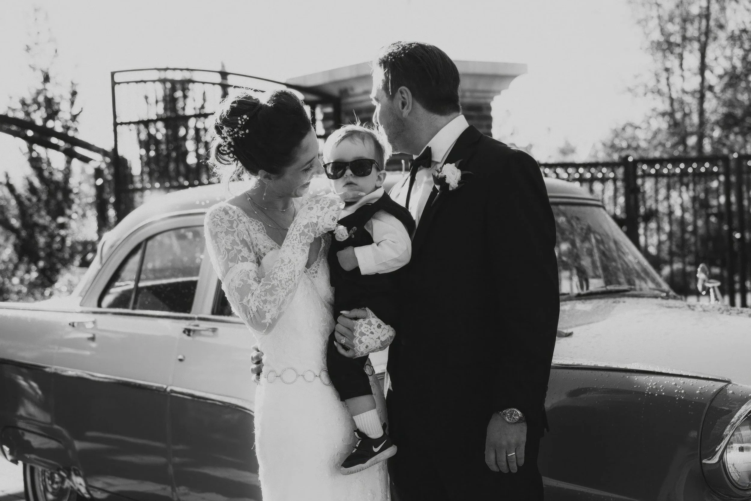 Bride and groom at a wedding holding a young boy wearing sunglasses and a tuxedo, standing in front of a vintage car.