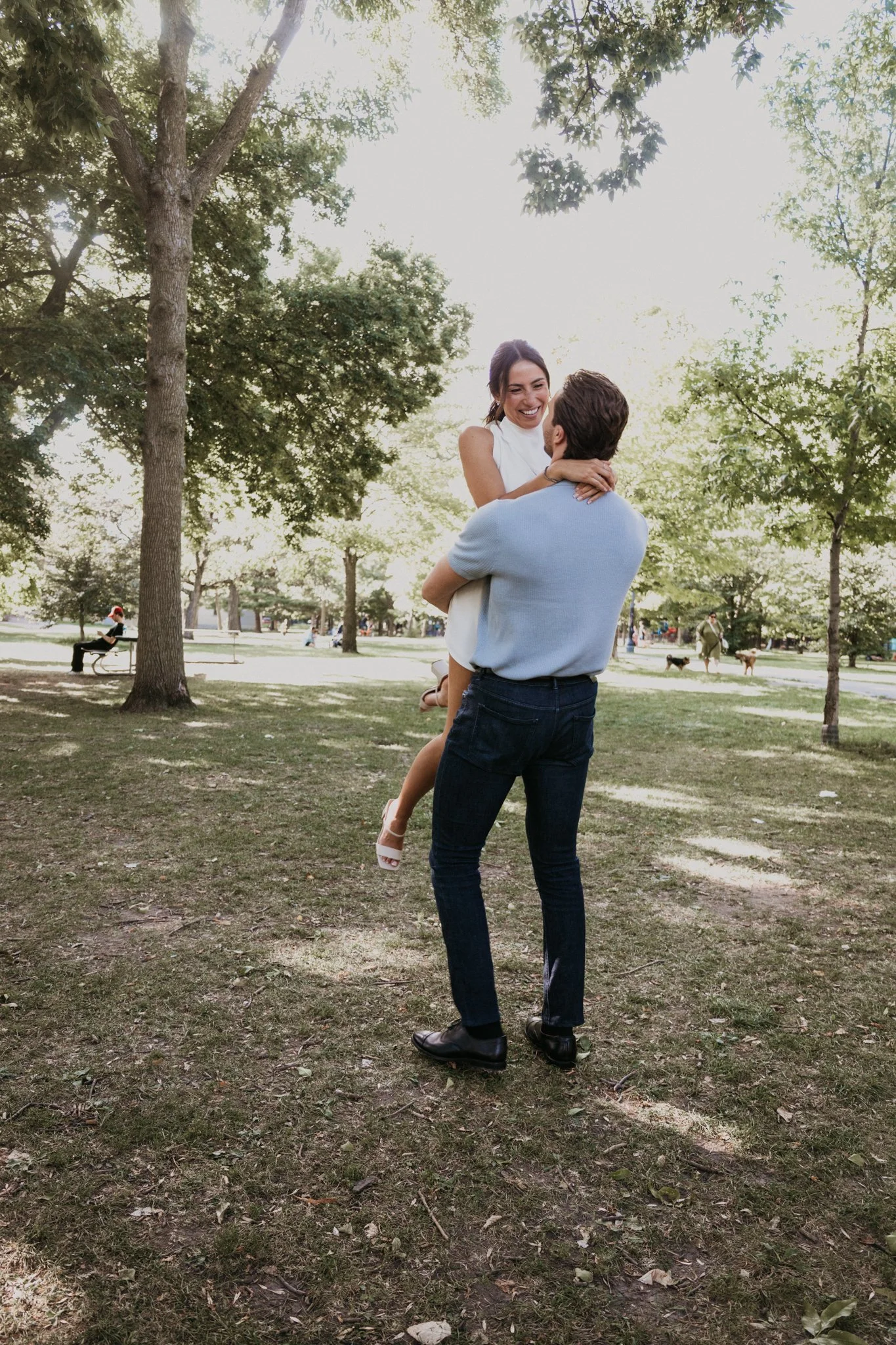 A couple in a park, the man holding the woman in his arms. The woman is smiling, and the scene is set among trees with other park visitors in the background.