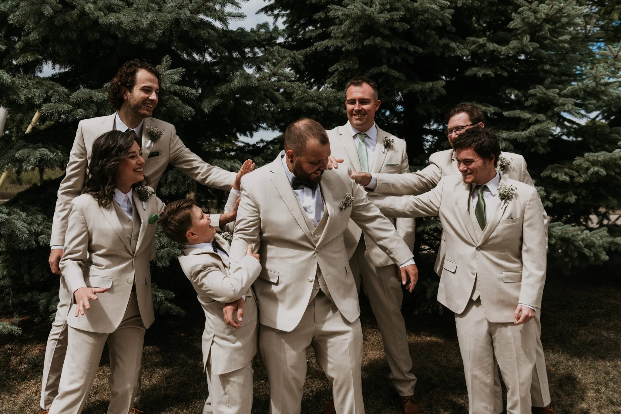 A group of seven men, dressed in light-colored suits, are gathered together outdoors in front of evergreen trees. They are smiling, laughing, and playfully touching or pointing at another man in the center with a beard and a bow tie. The scene appear