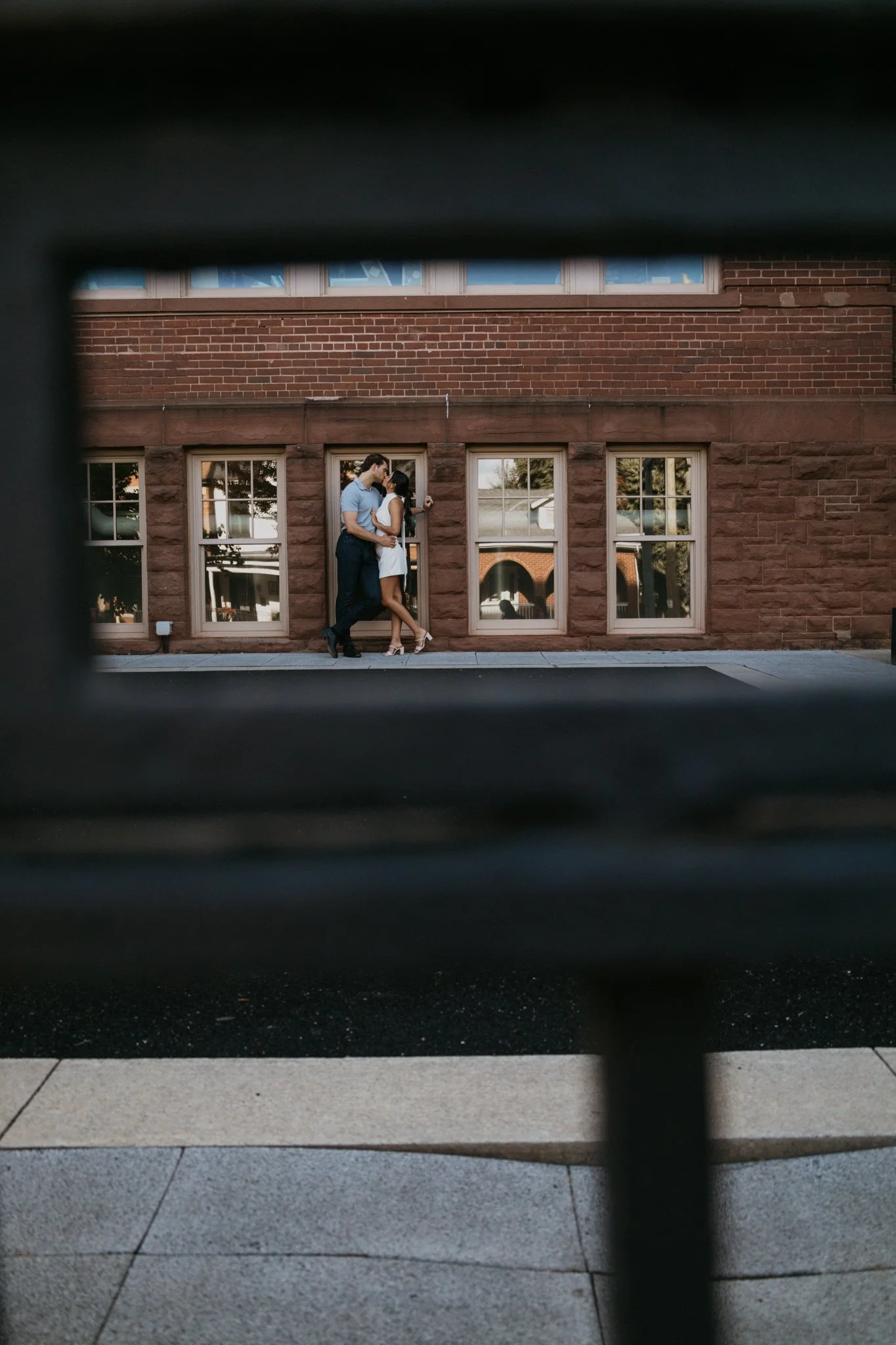 Two people kissing on a sidewalk behind a brick building, seen through a window or gap.