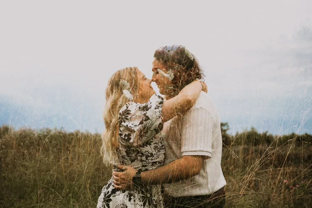 A couple standing close together in a grassy field, touching noses and gazing into each other's eyes.