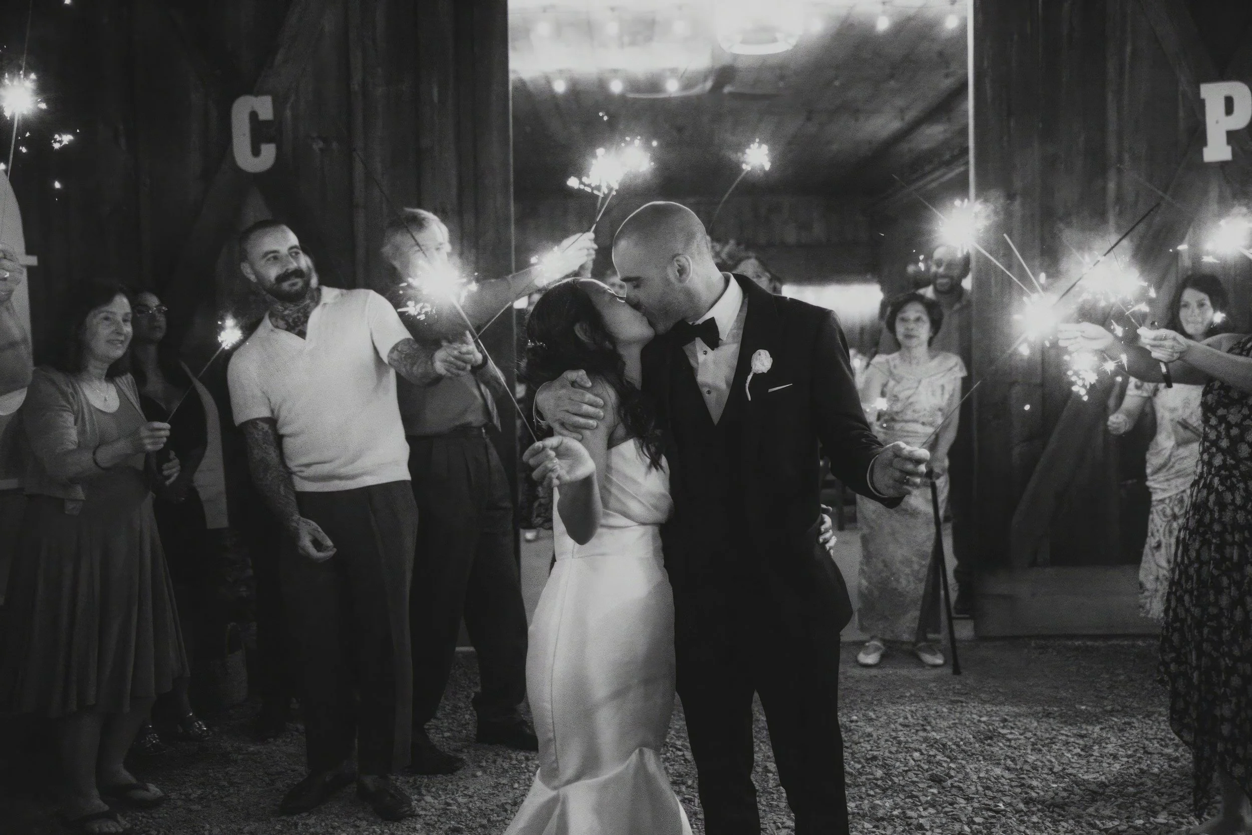 A newlywed couple shares a kiss at their wedding reception, surrounded by guests holding sparklers in a rustic indoor setting.