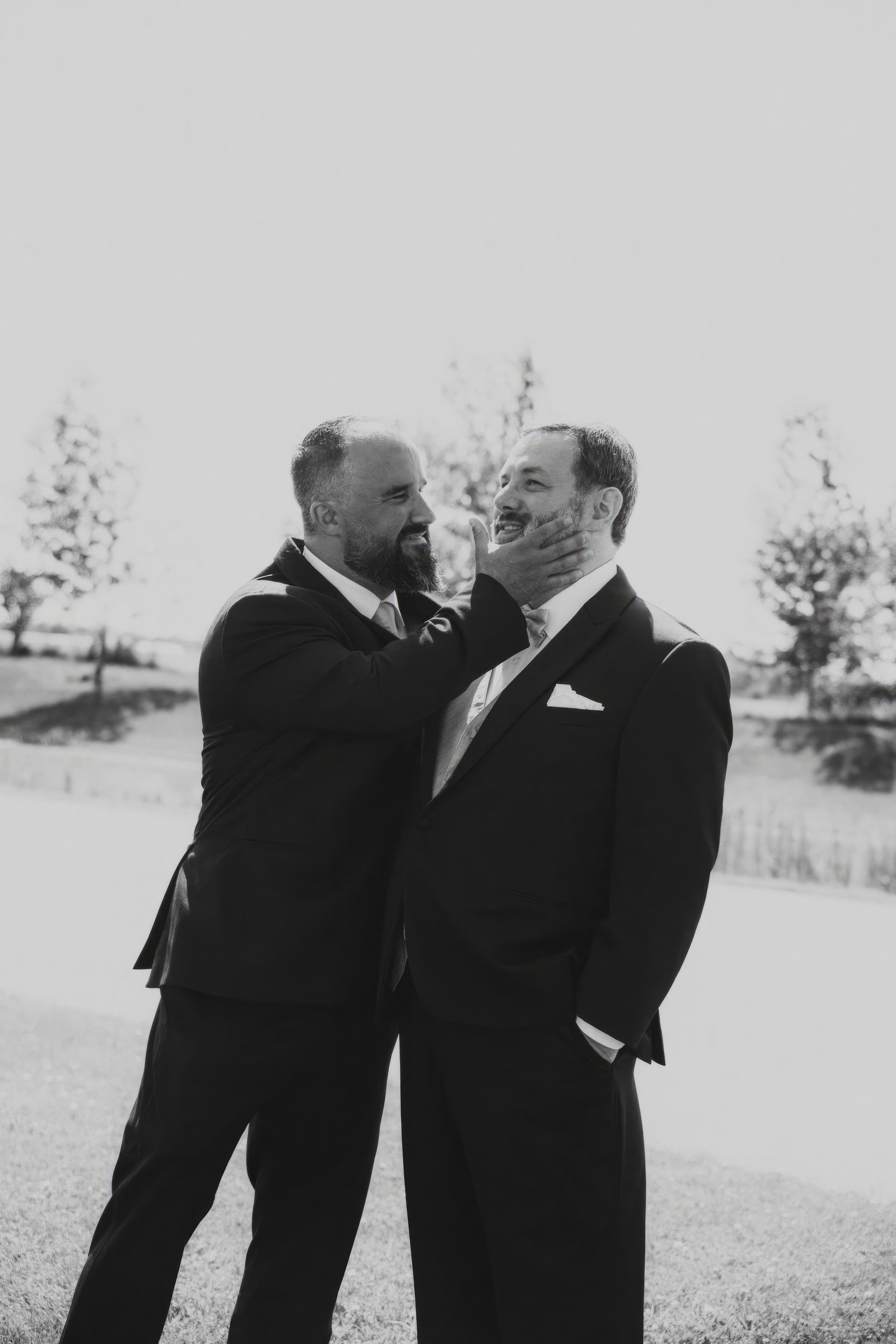 Two men in suits, one with a beard, smiling and playfully touching the other's chin outdoors with trees and grass in the background.