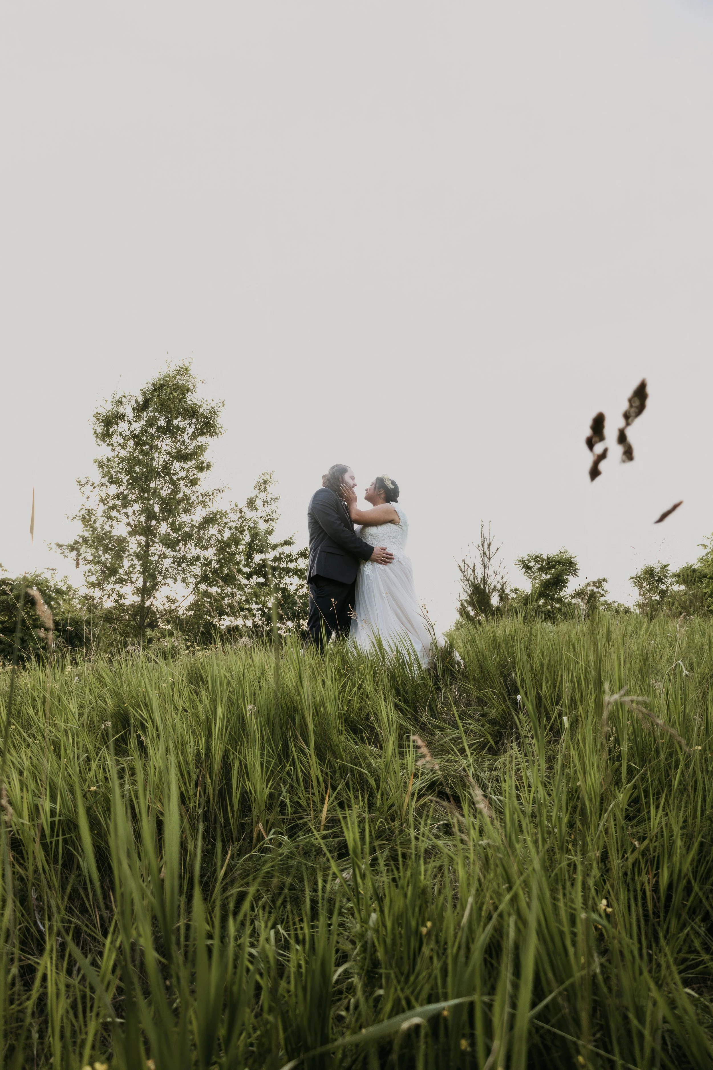 A bride and groom sharing a kiss in a grassy field with trees in the background during sunset.