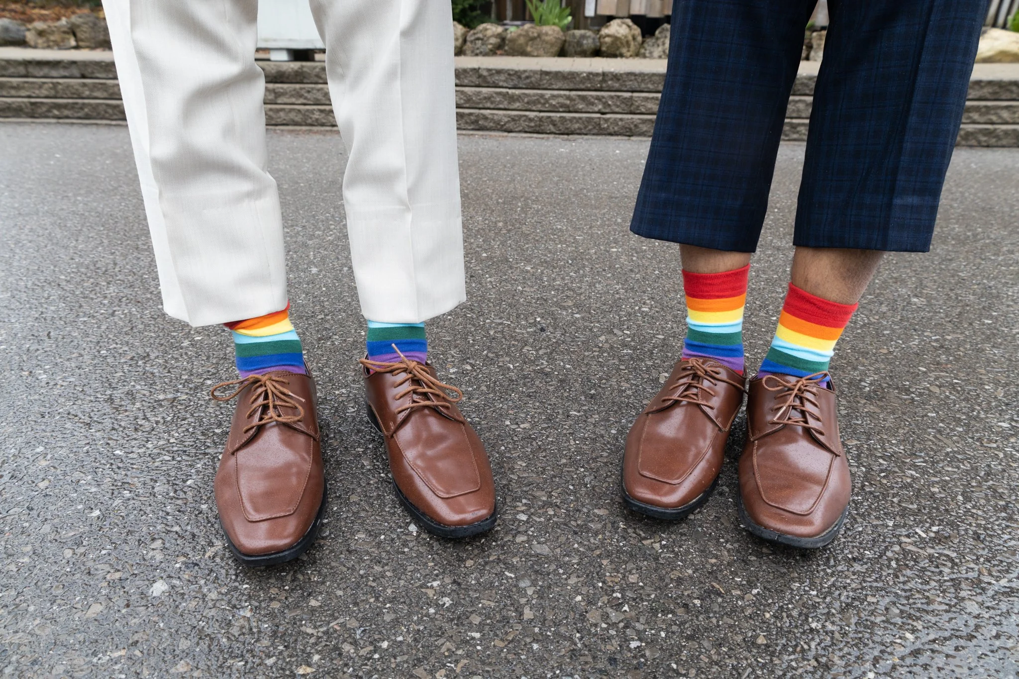 Two people wearing brown shoes and rainbow-colored socks standing on pavement, with one person in white pants and the other in blue plaid pants.