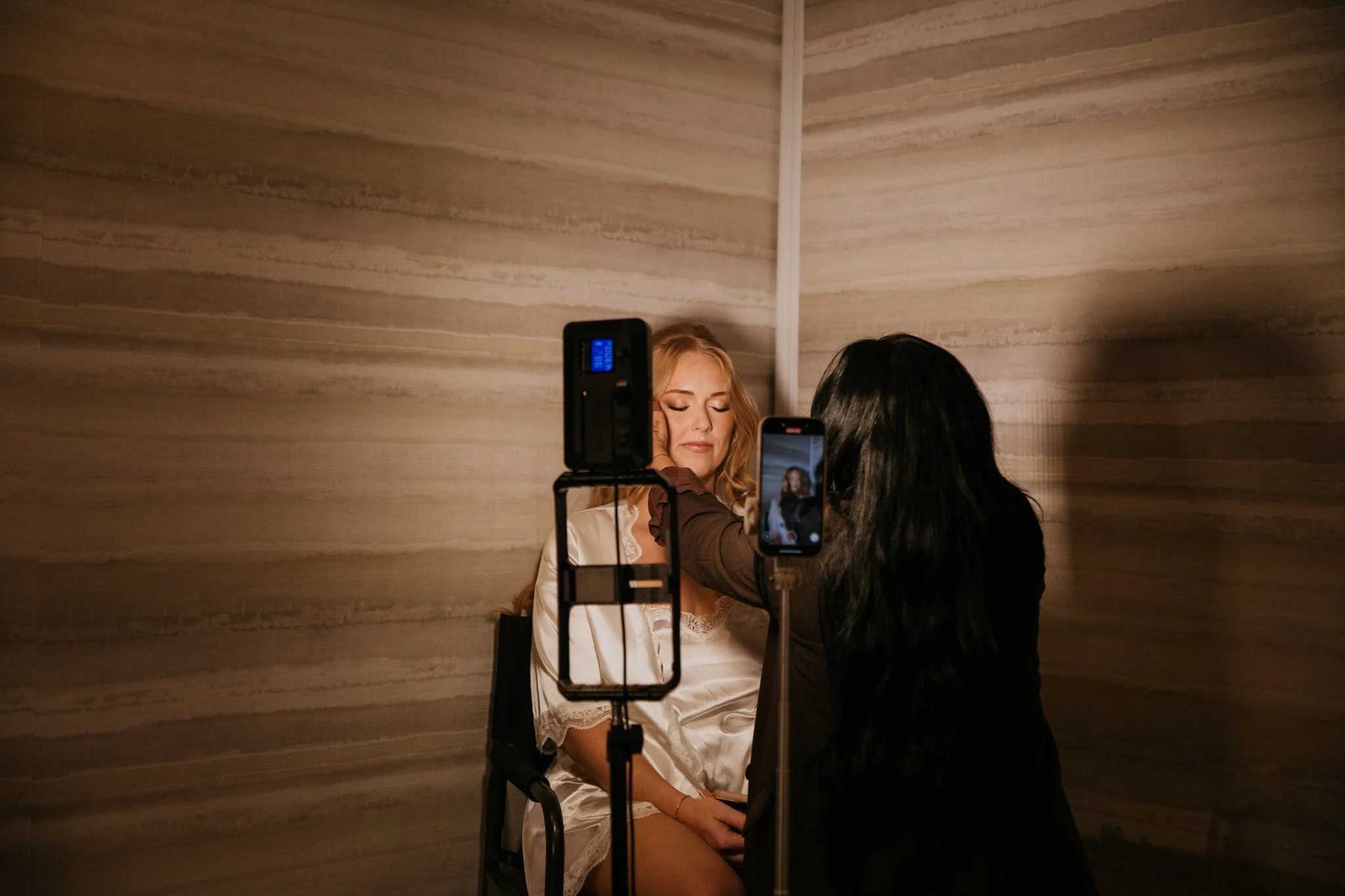 A woman with blonde hair and a satin robe sitting with eyes closed while a makeup artist applies makeup to her face, with two smartphones filming the scene, one on each side.