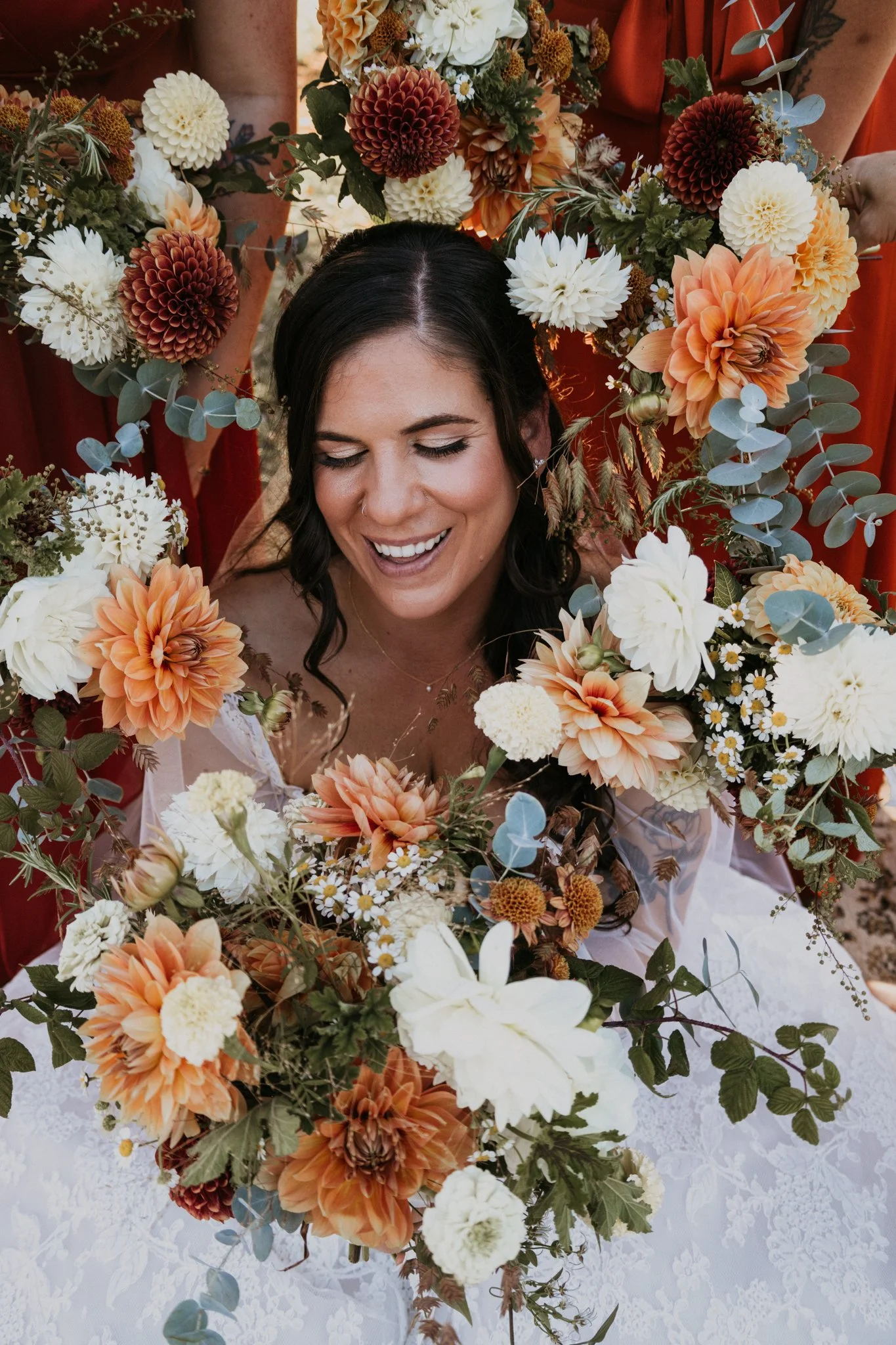A woman with dark hair, smiling and looking down, surrounded by a large, colorful bouquet of flowers with shades of white, cream, peach, and deep red, with greenery and eucalyptus leaves. She appears to be at a wedding or celebration.