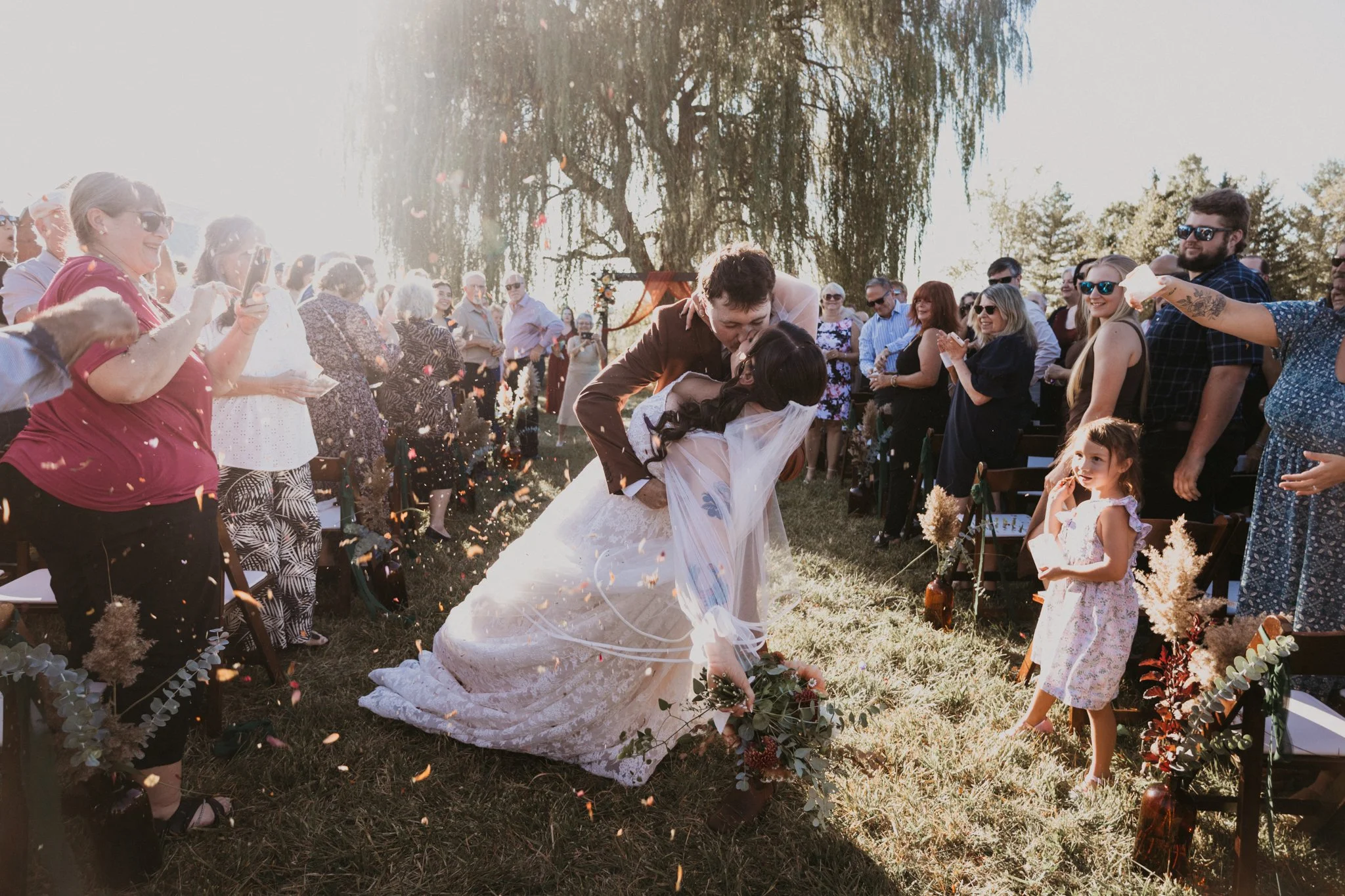 A bride and groom kiss outdoors at their wedding ceremony while guests celebrate and throw confetti.