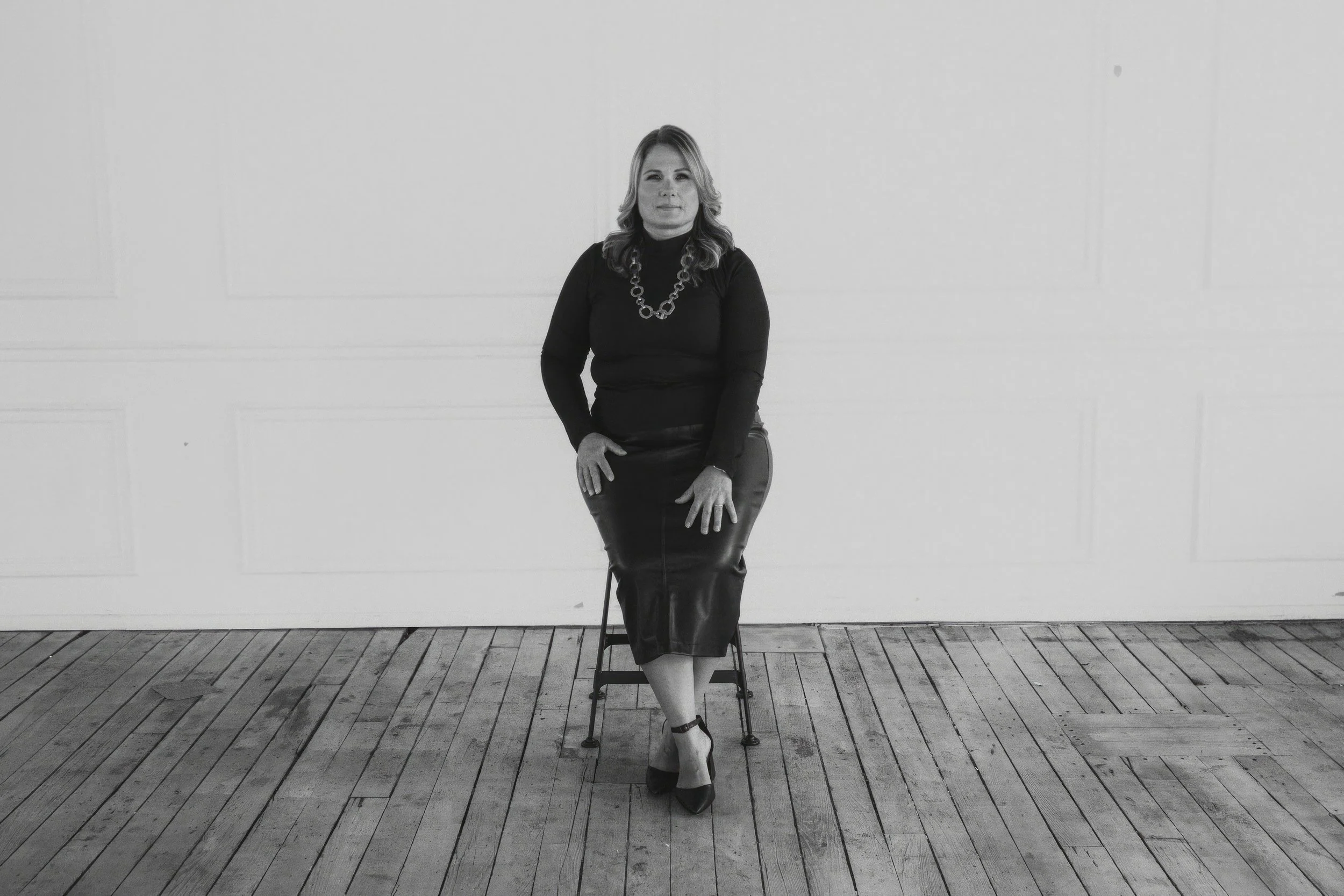 A woman with wavy hair sitting on a chair against a plain wall, dressed in a black turtleneck, a leather skirt, with a chunky chain necklace, and wearing heels, in a minimalistic setting with wooden floors.