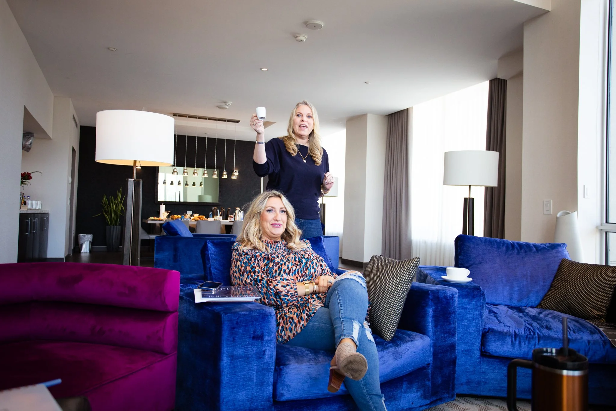 Two women in a hotel suite, one standing and speaking, the other sitting on a blue velvet couch, with a dining area in the background.