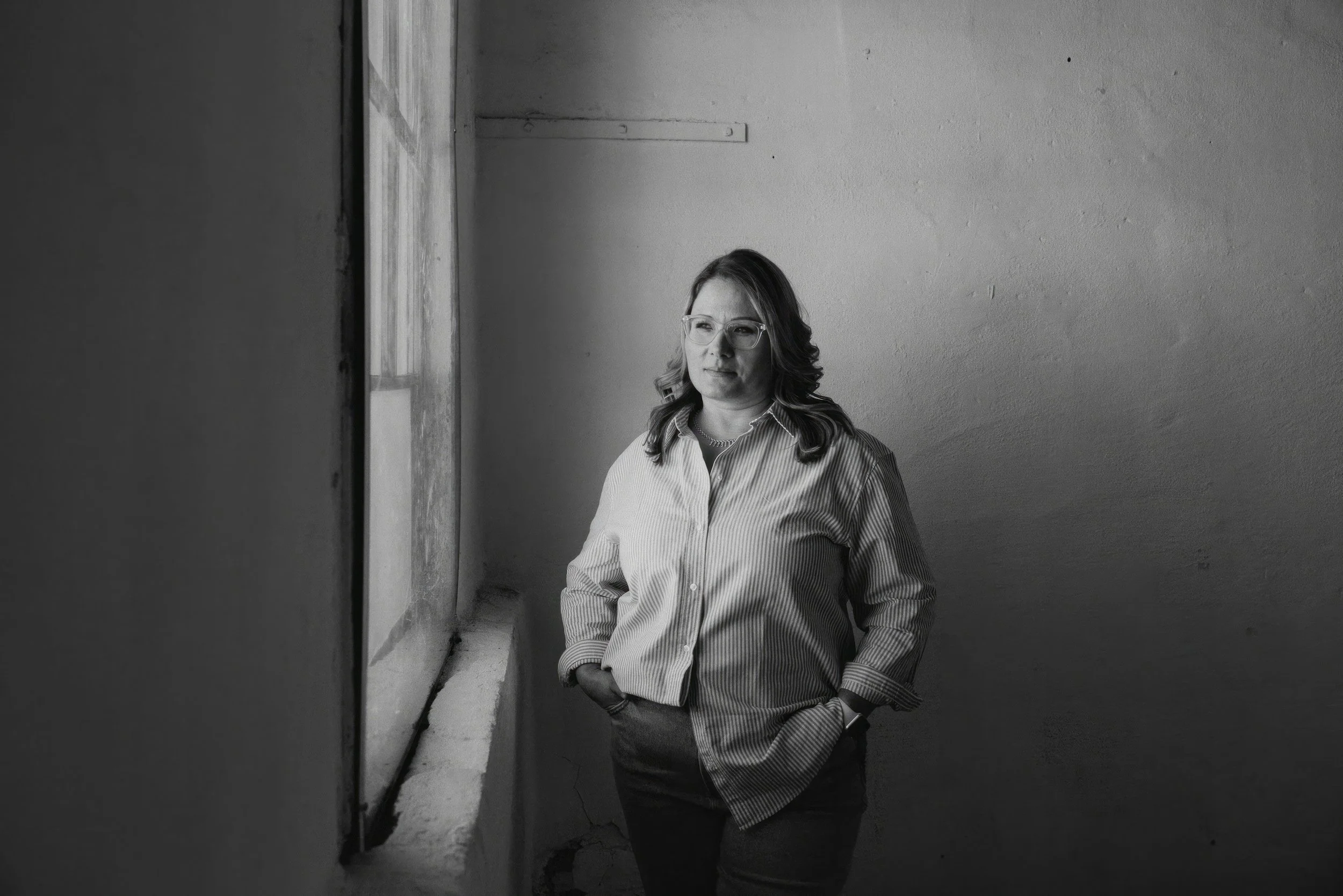 A woman with glasses and wavy hair standing indoors near a window with natural light, wearing a striped shirt and looking contemplative.
