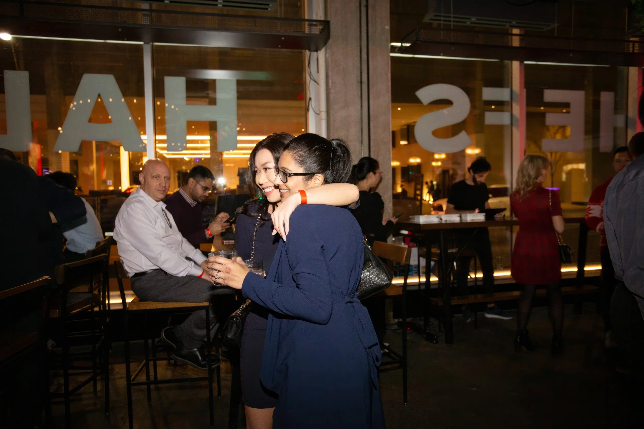 Two women hugging and smiling at a social gathering in a warmly lit indoor space with other people in the background.