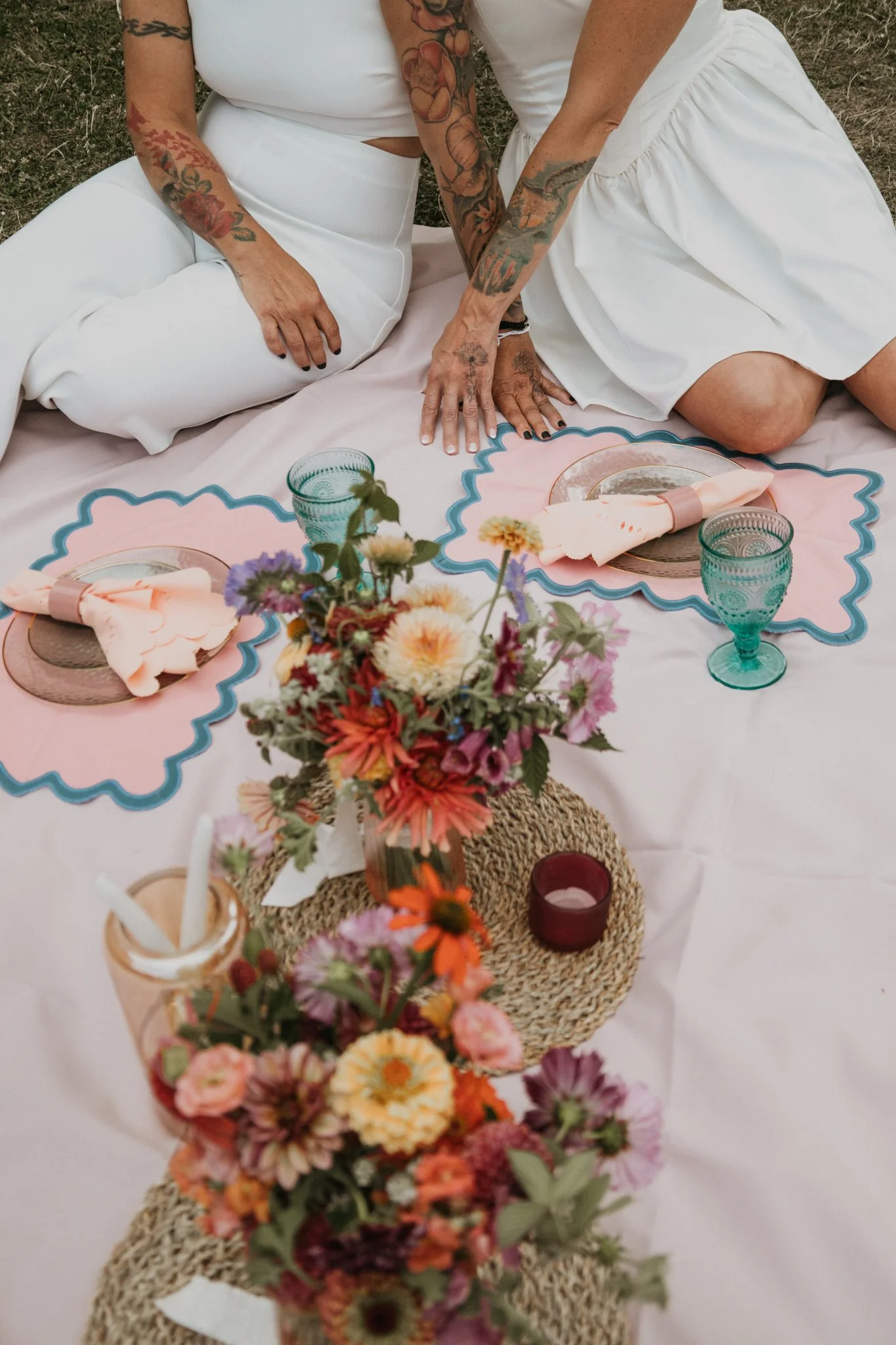 Two women with tattoos sit on a pink and blue decorated tablecloth during an outdoor gathering with a floral centerpiece, drink glasses, and tableware.