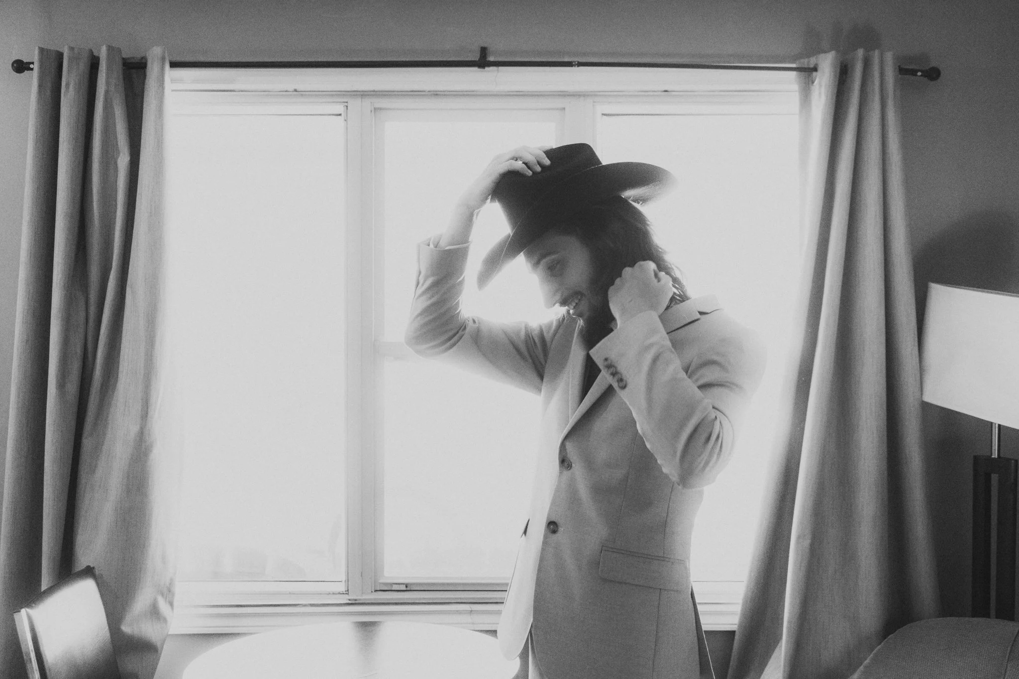 A man wearing a light-colored suit and a wide-brimmed hat, smiling, adjusting his hat with one hand and holding his neck with the other, standing in front of a window with curtains.