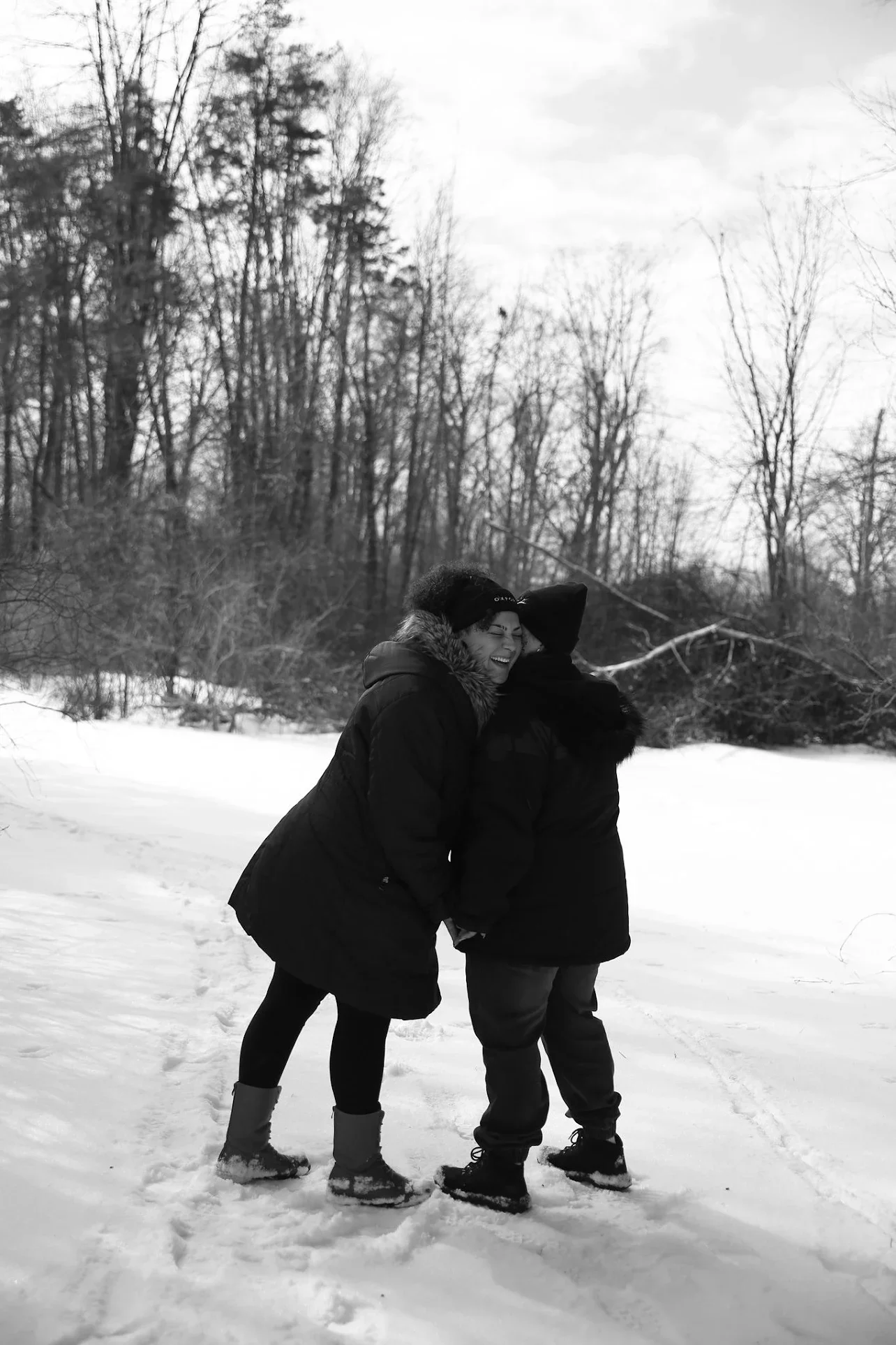 Two women in winter clothing laughing and hugging outdoors in a snow-covered landscape with trees in the background.