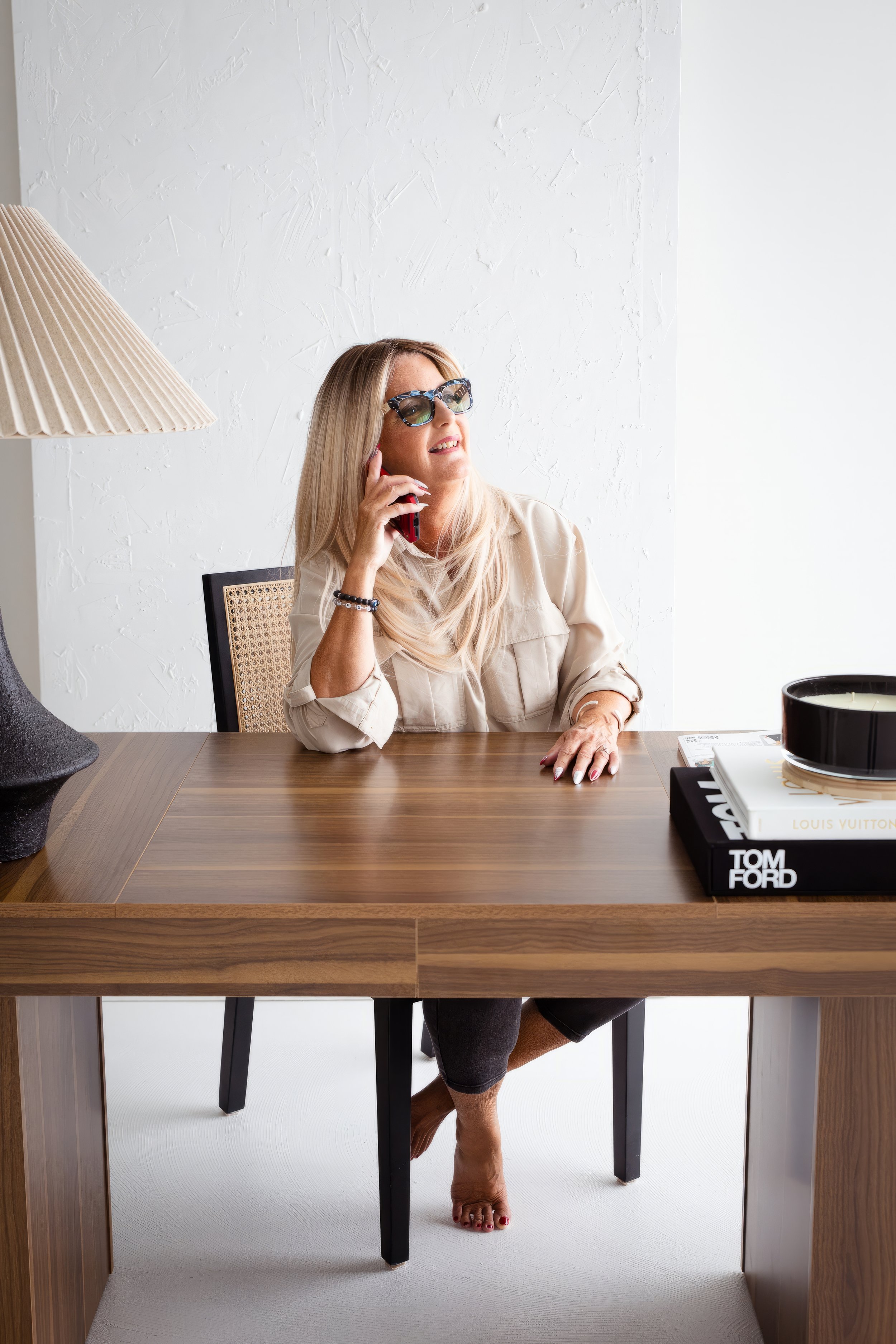 A woman sitting at a wooden desk, talking on a cellphone, wearing sunglasses, with books and a candle in a black tray on the desk. The room has a white textured wall and a lamp to her left.
