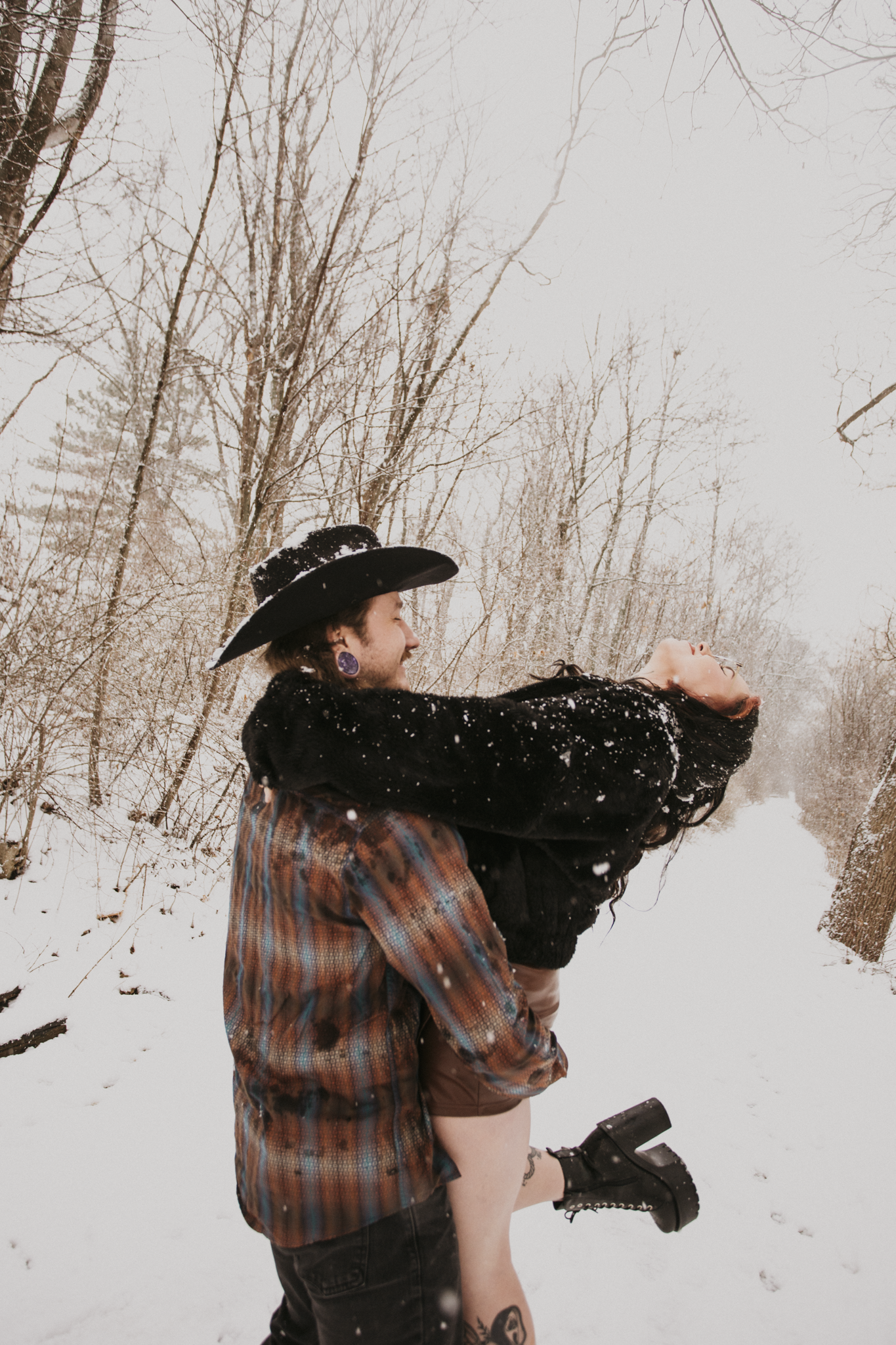 A person in a cowboy hat and plaid shirt holding a woman in black with long hair and black shoes in a snowy outdoor setting.