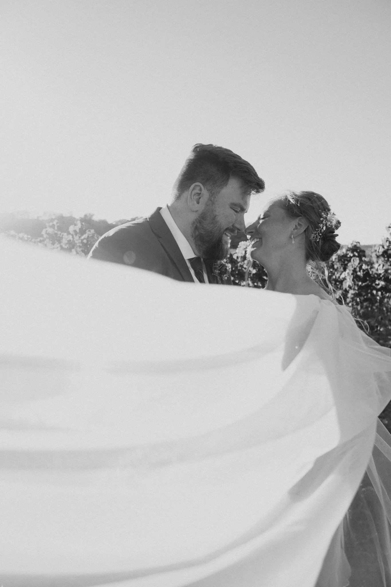 Black and white photo of a bride and groom smiling close together outdoors, with the bride's flowing veil partially covering them.