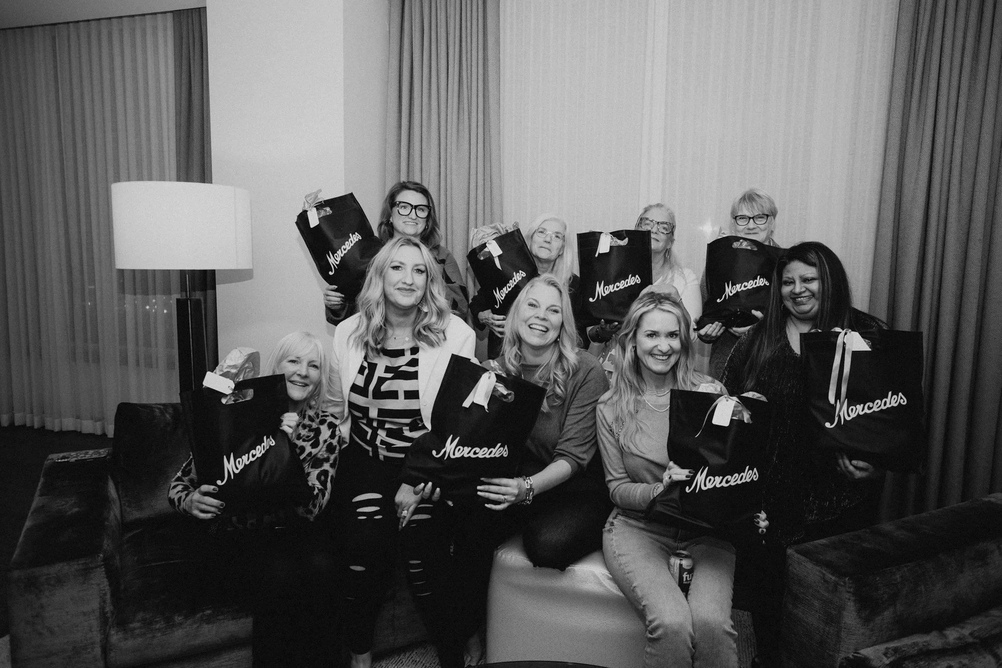 Group of ten women sitting and standing in a living room holding Mercedes-branded gift bags, smiling for a photo, in black and white.