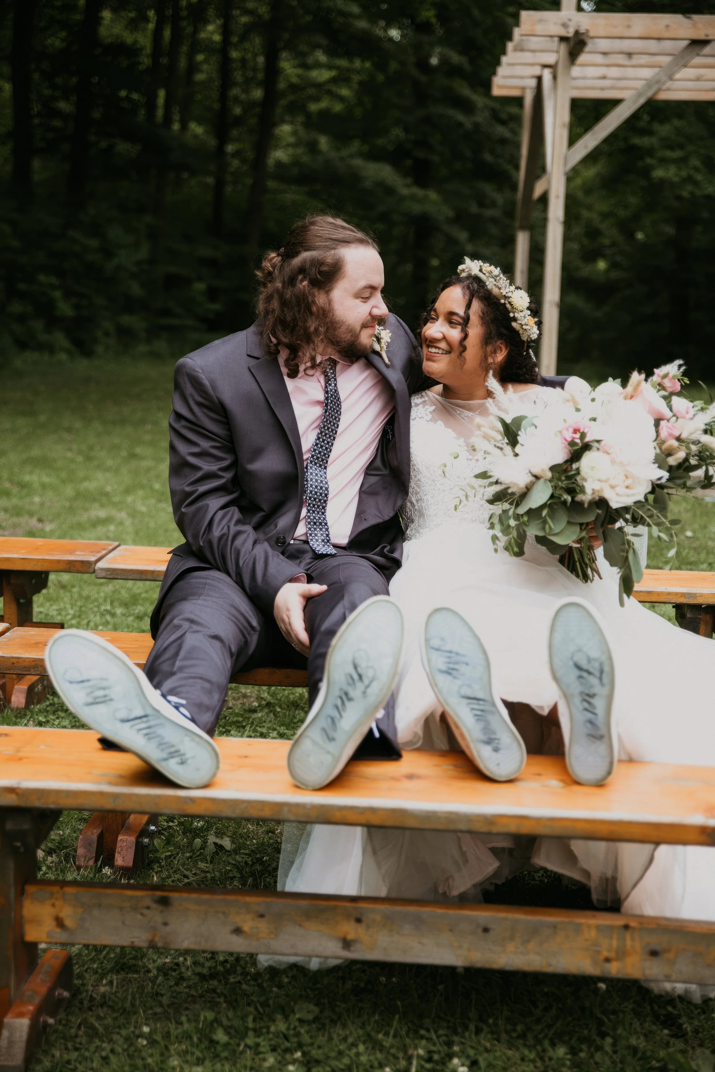 A newlywed couple sitting on a bench outdoors, smiling and looking at each other, with their shoes visible showing the words 'Happily Ever After' written on the soles. The bride holds a large bouquet of flowers, and they are surrounded by a green, wo