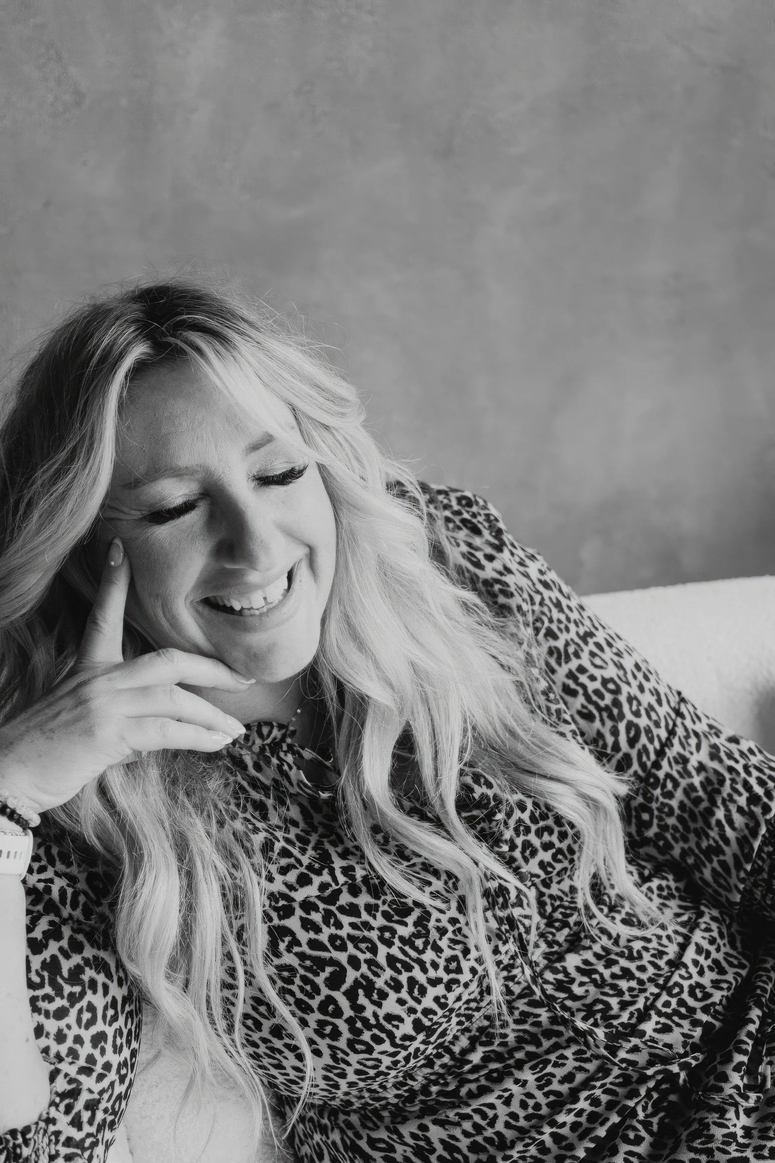 Black and white photo of a woman with long blonde hair smiling, wearing a leopard print shirt, sitting against a plain wall.