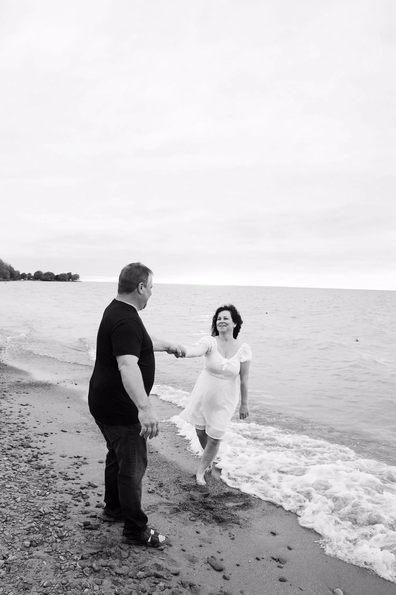 A man and woman holding hands on a beach, with waves reaching their feet, smiling and enjoying each other's company.