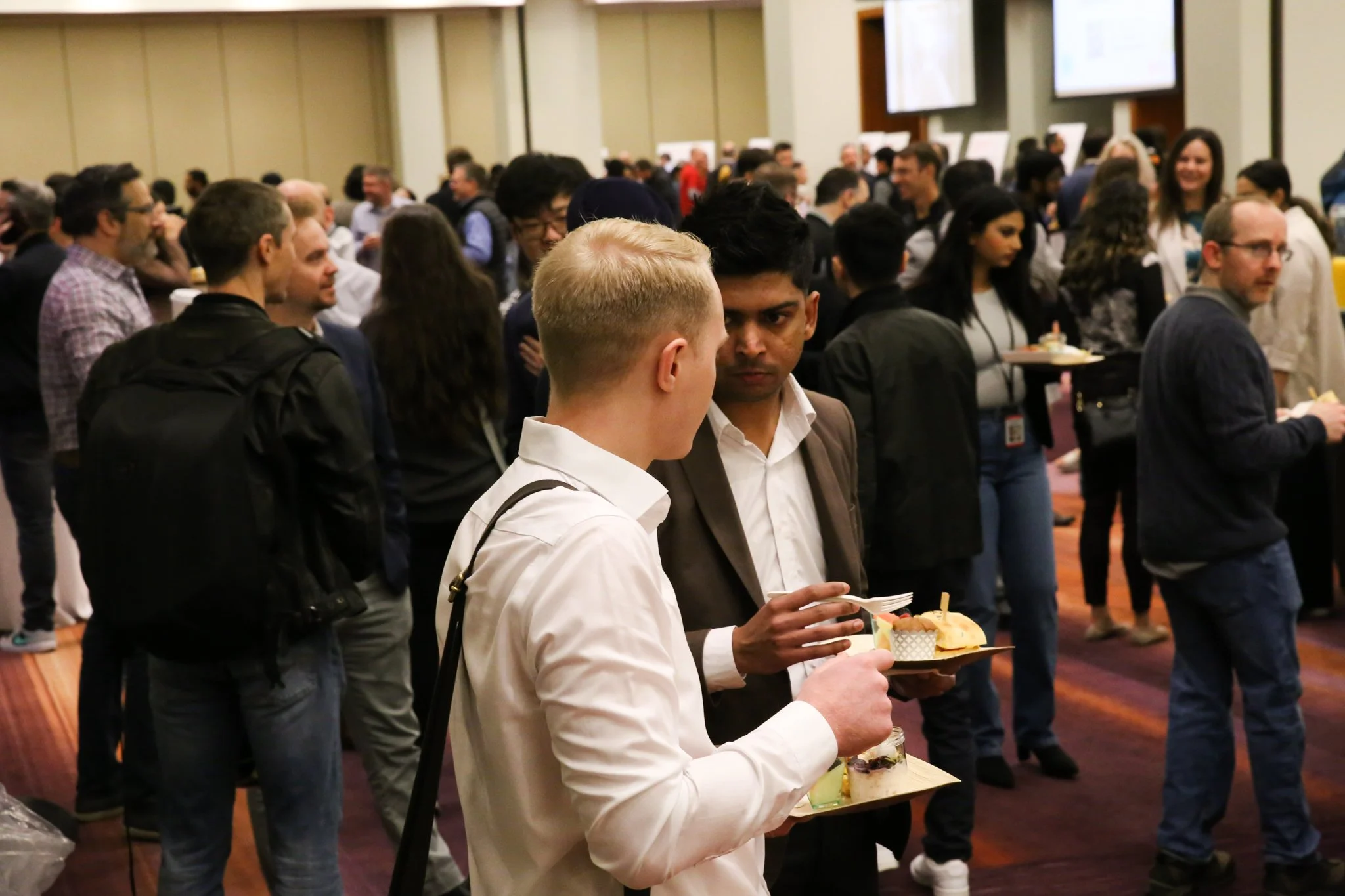 Two men in business attire eating at a crowded conference or networking event, with many people in the background