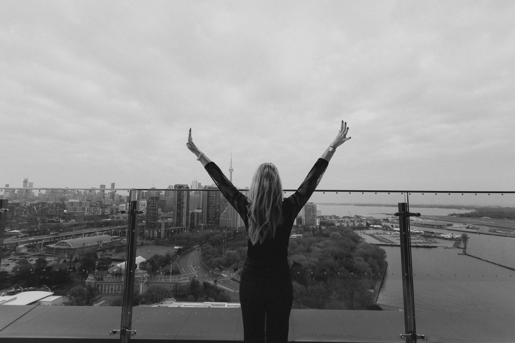 A woman with long hair, wearing a dark top, stands on a rooftop balcony with her arms raised in a 'V' shape, overlooking a city skyline including the CN Tower in Toronto, Canada, with a river and harbor in the background.