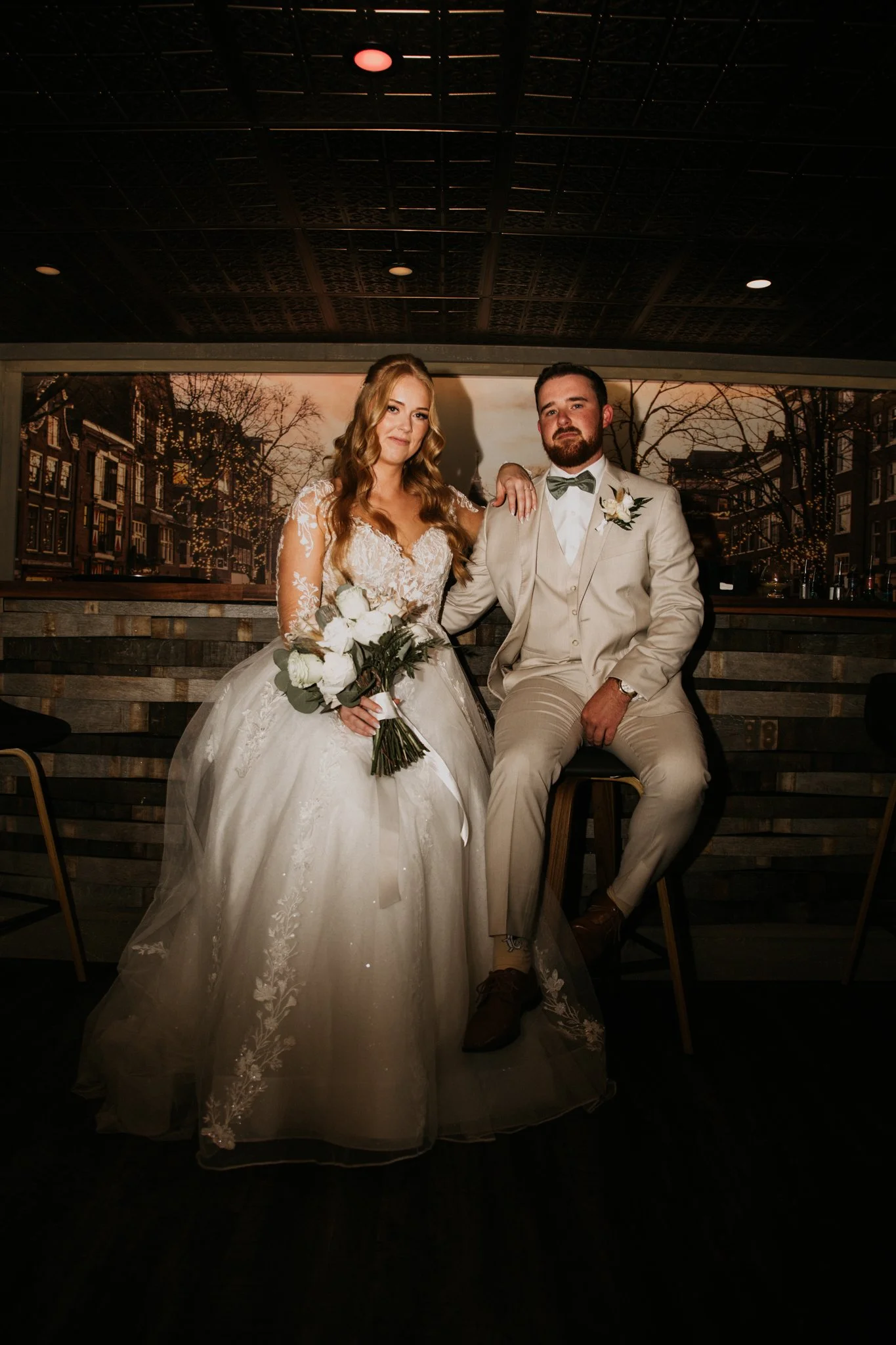 A bride in a white wedding gown holding a bouquet and a groom in a beige suit with a bow tie sitting on a high stool at an indoor wedding reception or venue.