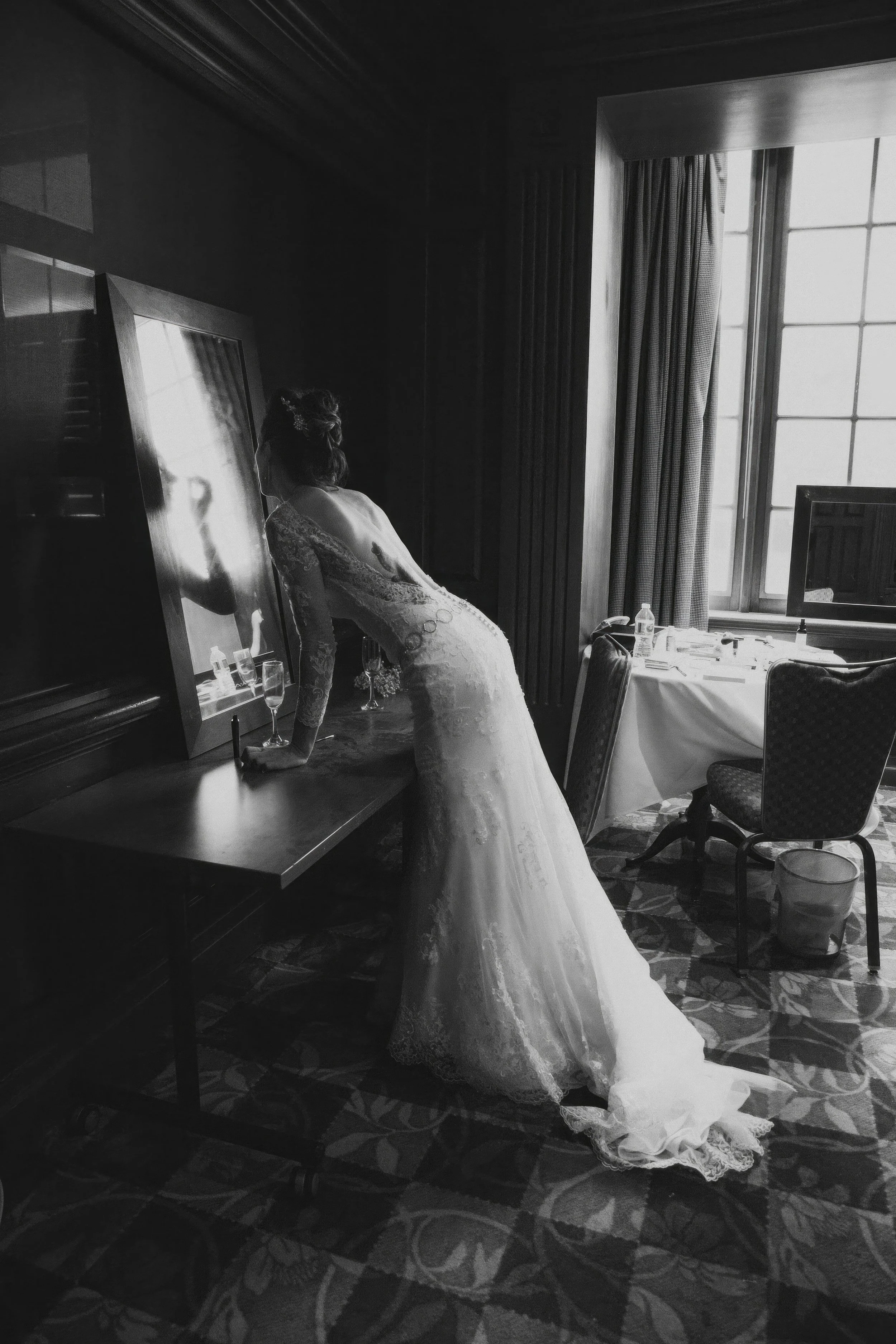 A bride in a lace wedding gown leaning over a table, looking into a mirror, with glasses and a water bottle nearby in a dimly lit room with a large window.